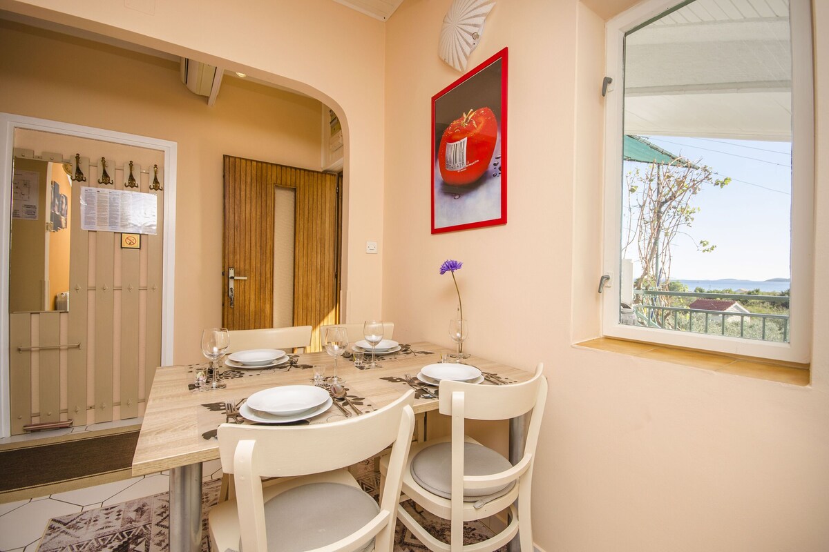Un coin repas avec une table dressée pour quatre personnes, des chaises blanches, une fenêtre donnant sur un beau paysage et un tableau encadré au mur.