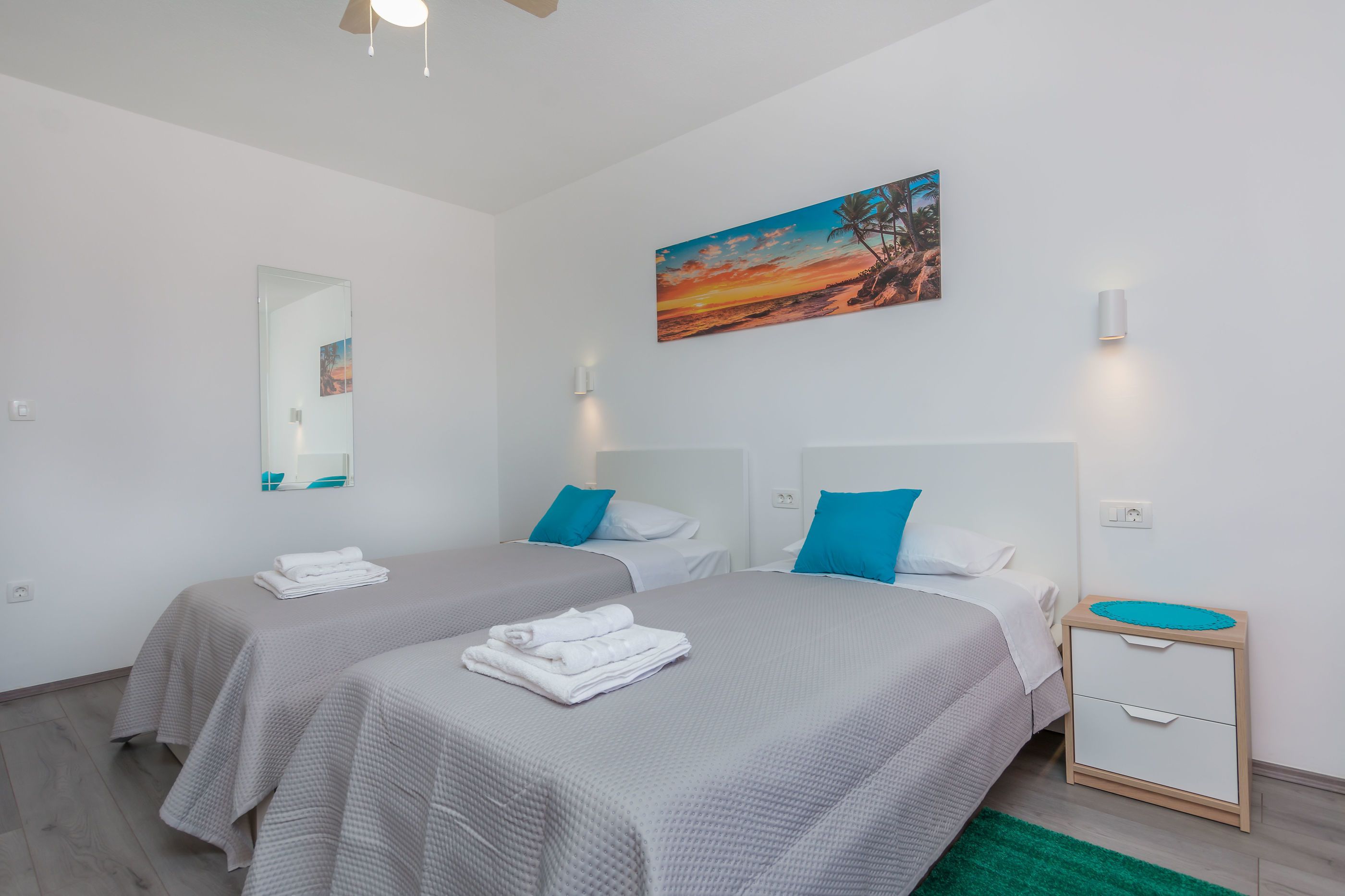 A hotel room with two beds, a nightstand, a mirror, and a painting of a sunset.