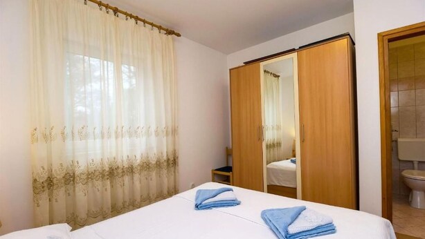 A hotel room with a bed, a wooden wardrobe, a bathroom with a toilet, and a window with sheer curtains. - Apartments Marlen (Mali Losinj)