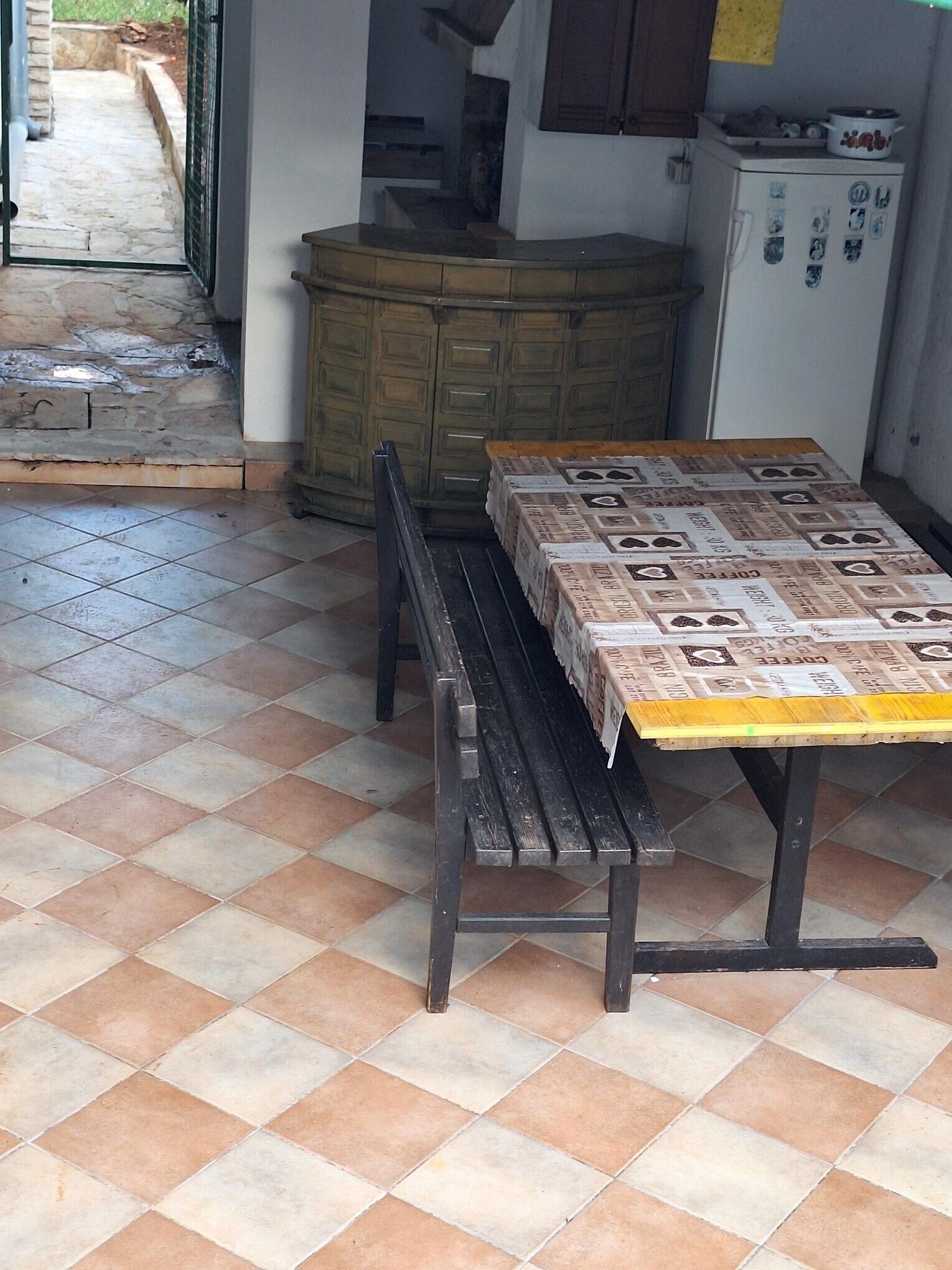 Uma cozinha com piso de azulejos, um banco de madeira, uma mesa com jornal e uma geladeira.