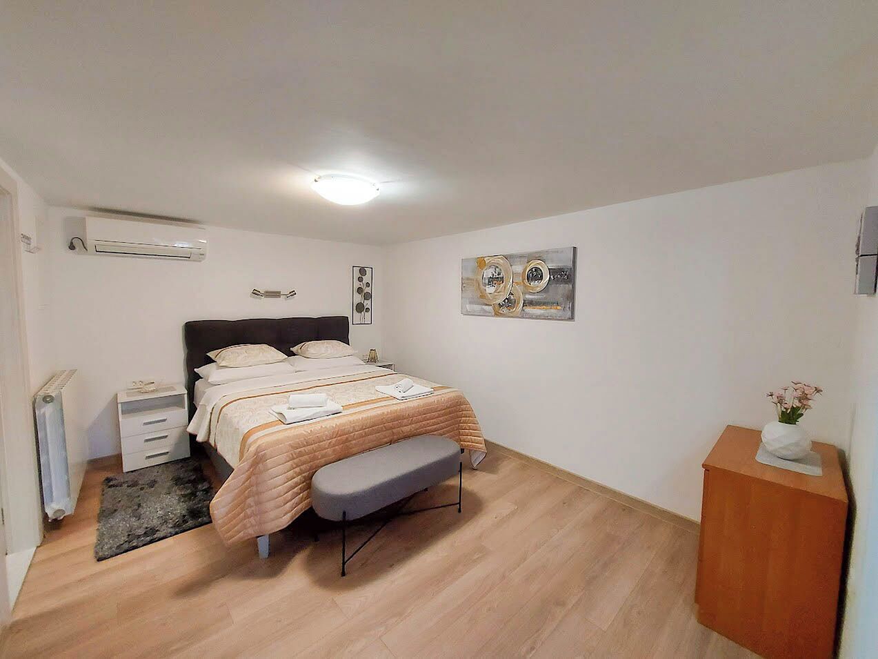 A bedroom with a bed, bedside tables, a radiator, a small rug, a bench, and a wall-mounted shelf with flowers.