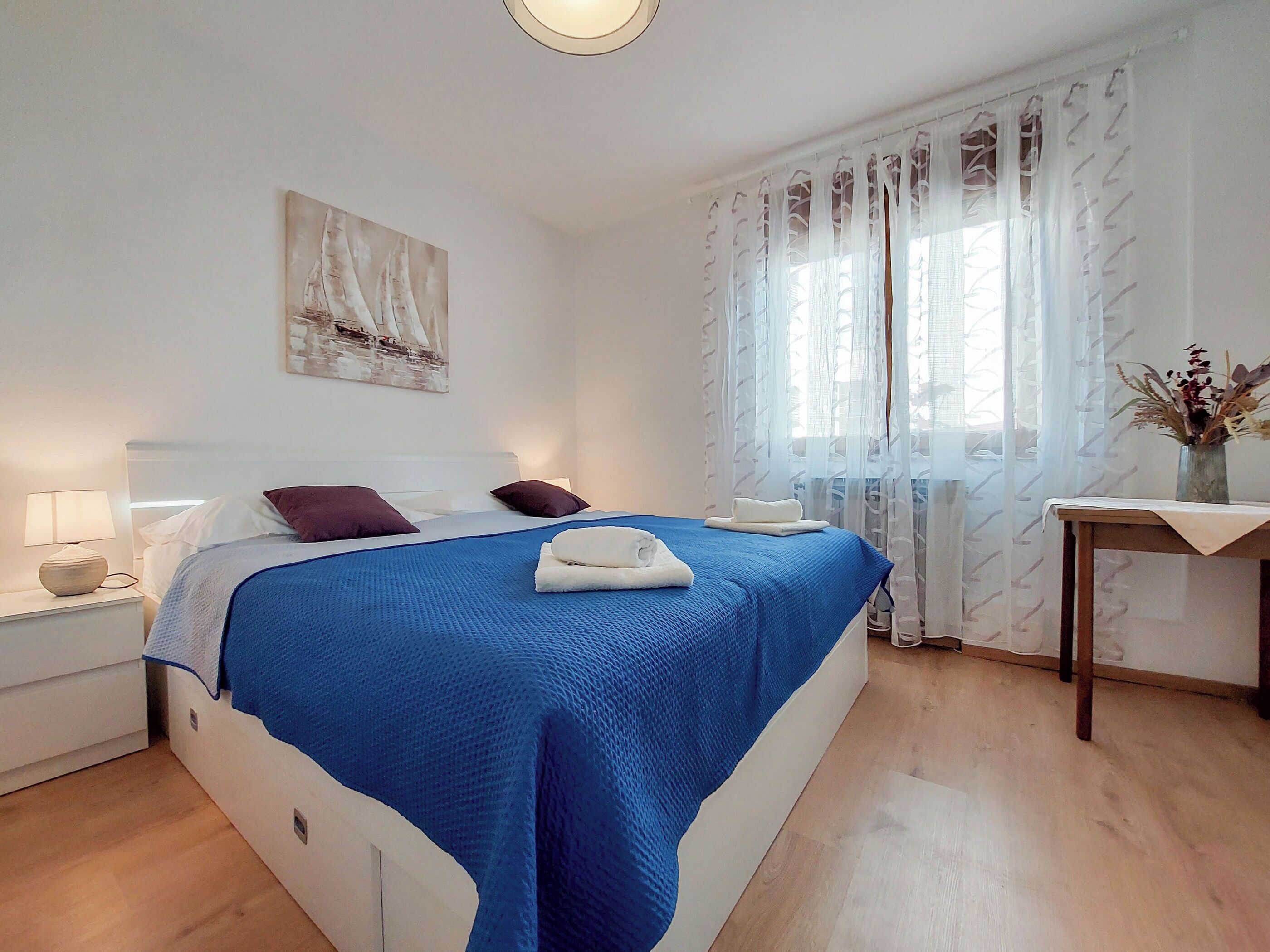 Une chambre à coucher avec un lit, une table de chevet, une lampe, un tableau, une fenêtre avec des rideaux légers et une petite table sur laquelle est posé un vase.