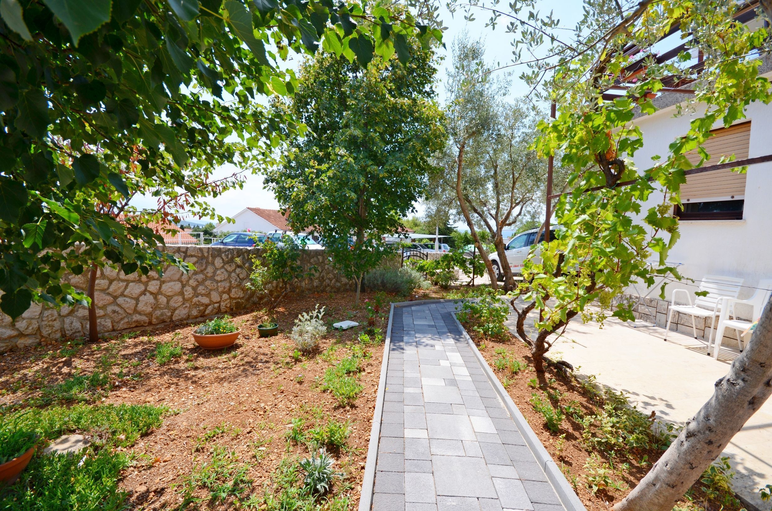 Un chemin pavé traverse un jardin où l’on trouve diverses plantes et arbres, le tout bordé d’un mur de pierre et d’un bâtiment blanc doté d’un balcon.