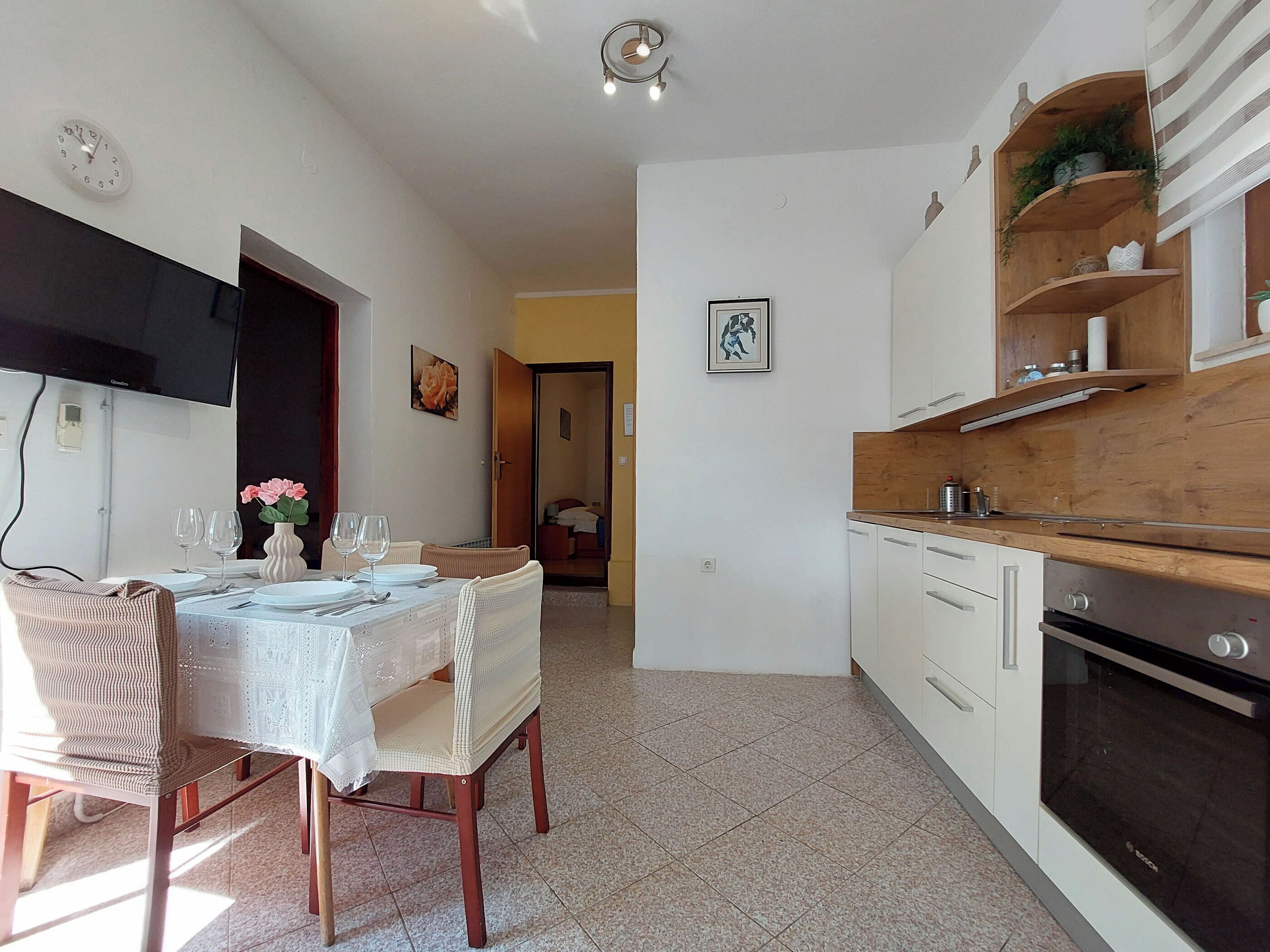 A compact kitchen with white cabinets, a wooden countertop, and a Bosch oven. There is a dining area with a table set for two and a chair.