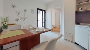 Un coin repas avec une table en bois, des chaises blanches et un vase rempli de fleurs. On y trouve également un espace cuisine avec des étagères et une porte qui donne sur une autre pièce.