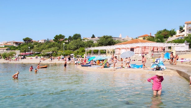 Une plage à l’eau claire, où l’on peut voir des gens nager et prendre des bains de soleil, et une tente blanche avec des auvents à rayures.
