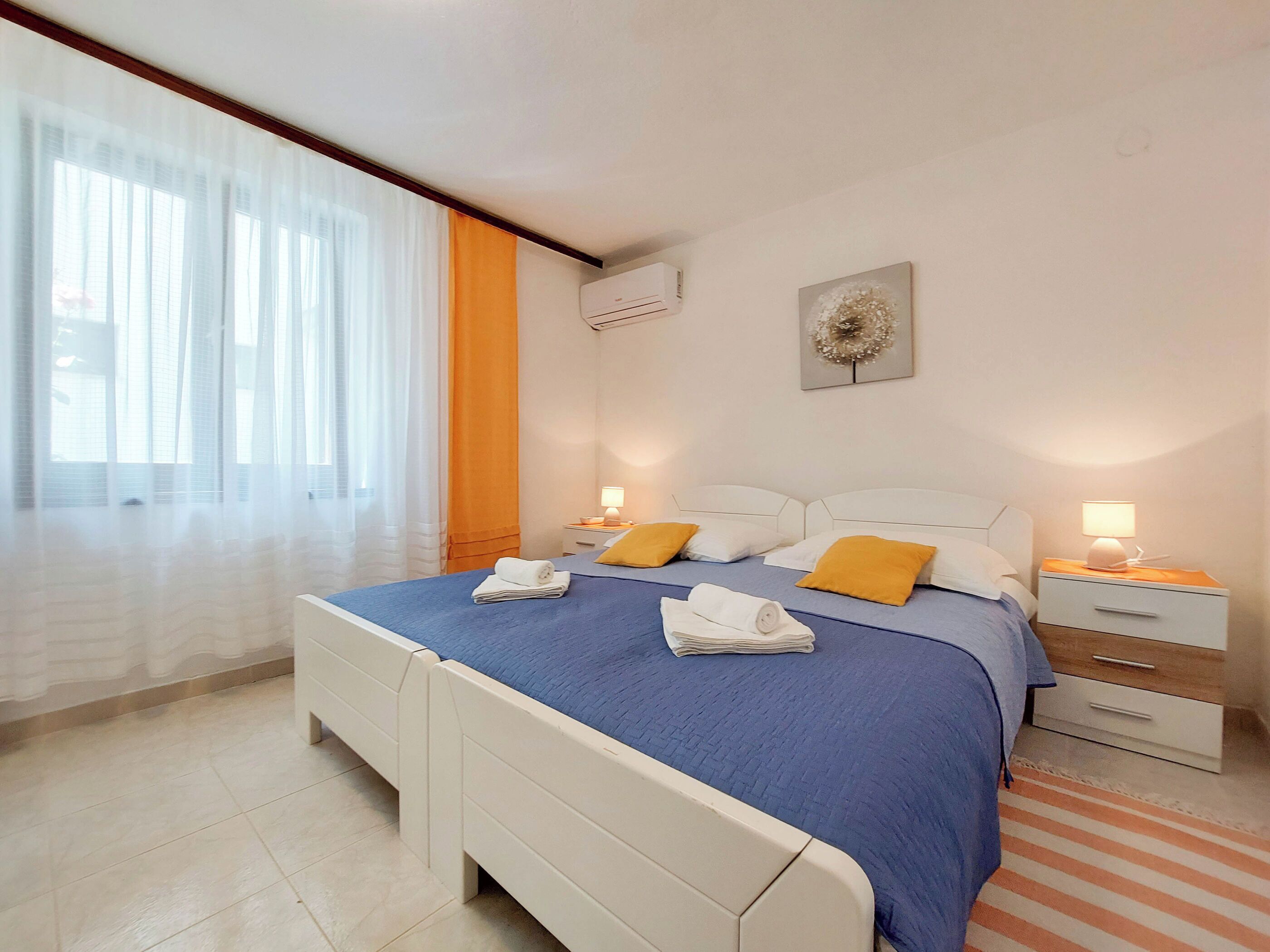 Une chambre d’hôtel avec un grand lit, deux tables de chevet, un climatiseur fixé au mur et une fenêtre avec des rideaux légers.