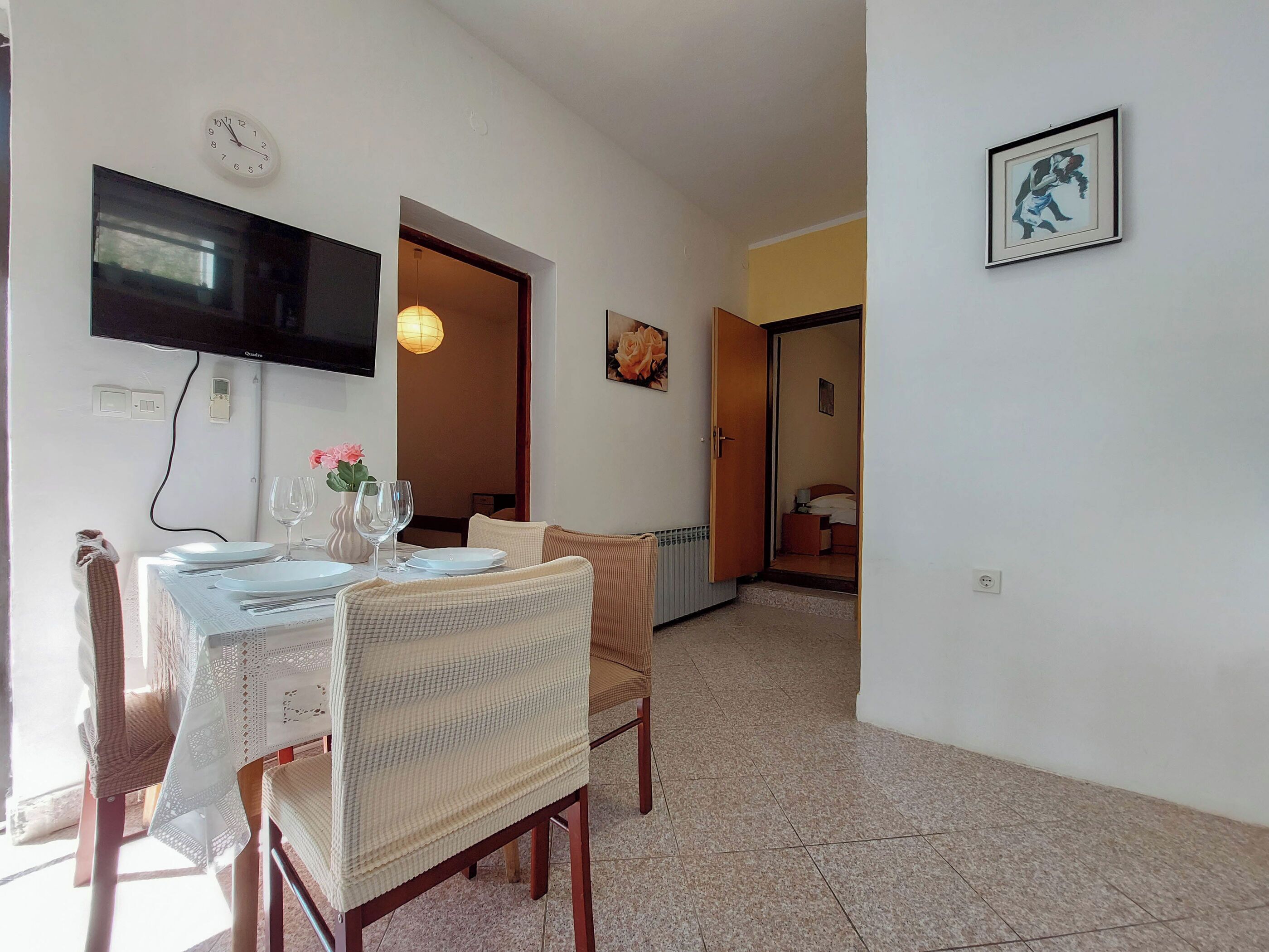 A dining area with a table set for two, a television mounted on the wall, and a framed picture on the wall.