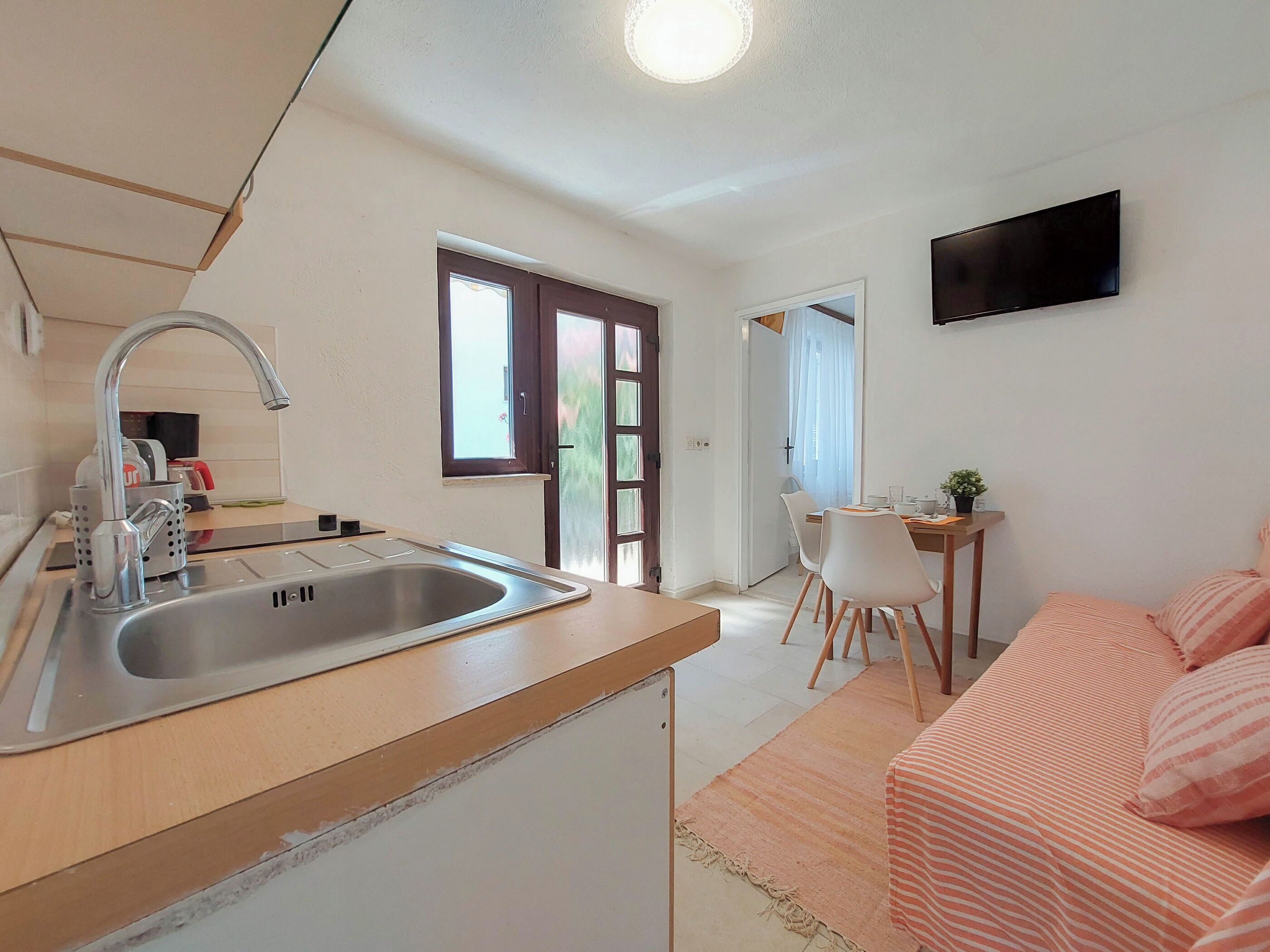 Une cuisine de taille réduite, équipée d’un évier, d’un coin repas avec une table et des chaises, d’un téléviseur fixé au mur et d’une porte donnant sur une autre pièce.