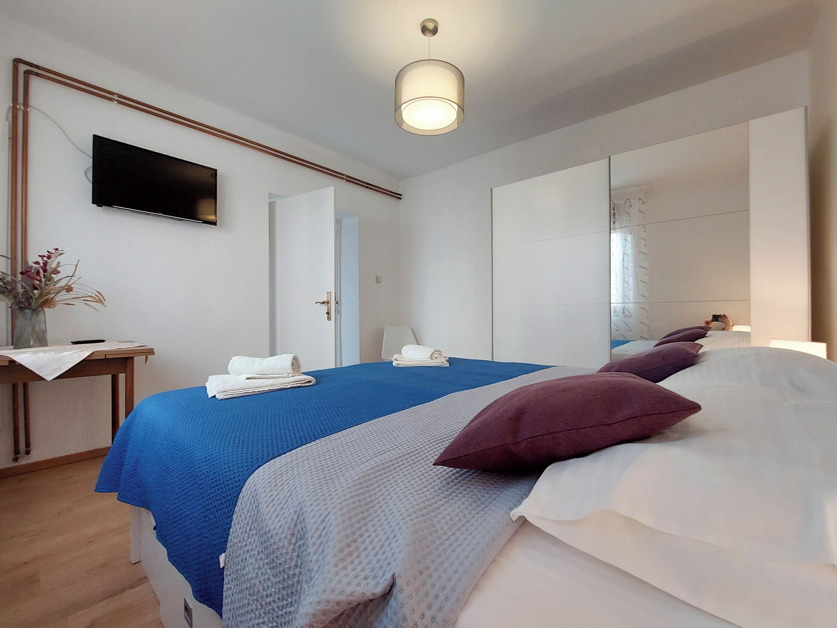 A modern hotel room with a large bed, a flat-screen TV, a desk with a vase of flowers, and a large mirror.