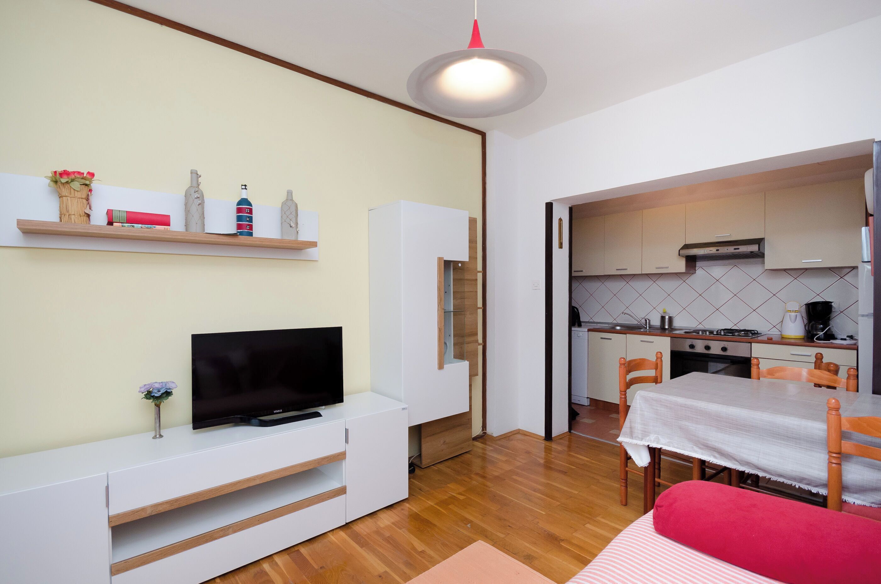 A compact living space with a TV on a white entertainment unit, a dining table with chairs, and a kitchen area with cabinets and appliances.