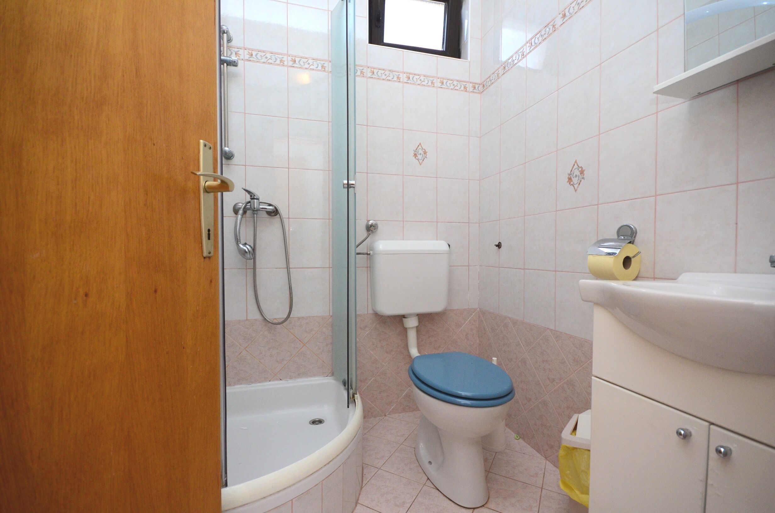 Ein Badezimmer mit Duschkabine aus Glas, Toilette mit blauem Sitz und Waschbecken mit Spiegel.