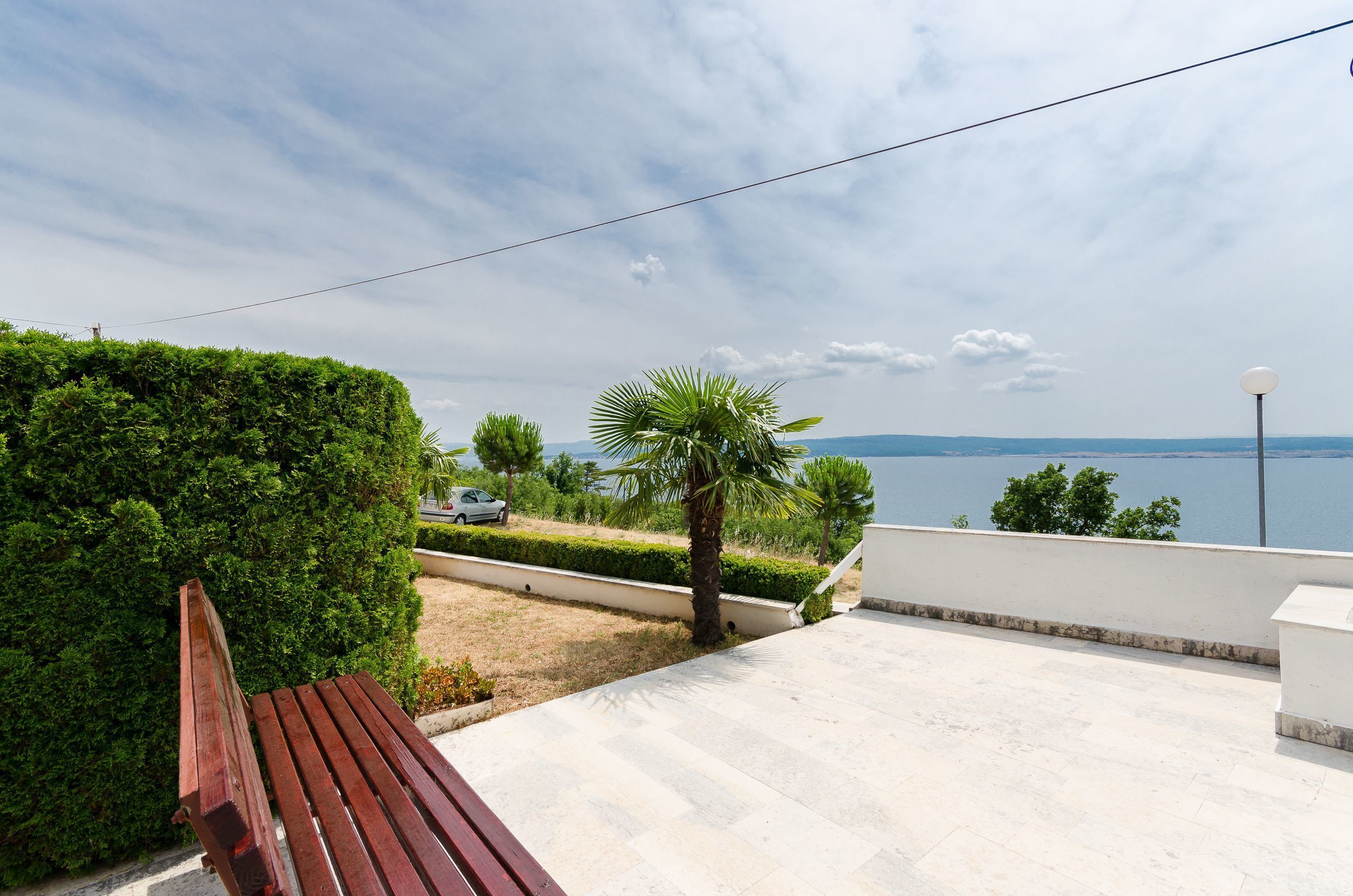 A terrace with a wooden bench, a view of a body of water, and a car parked in the background.
