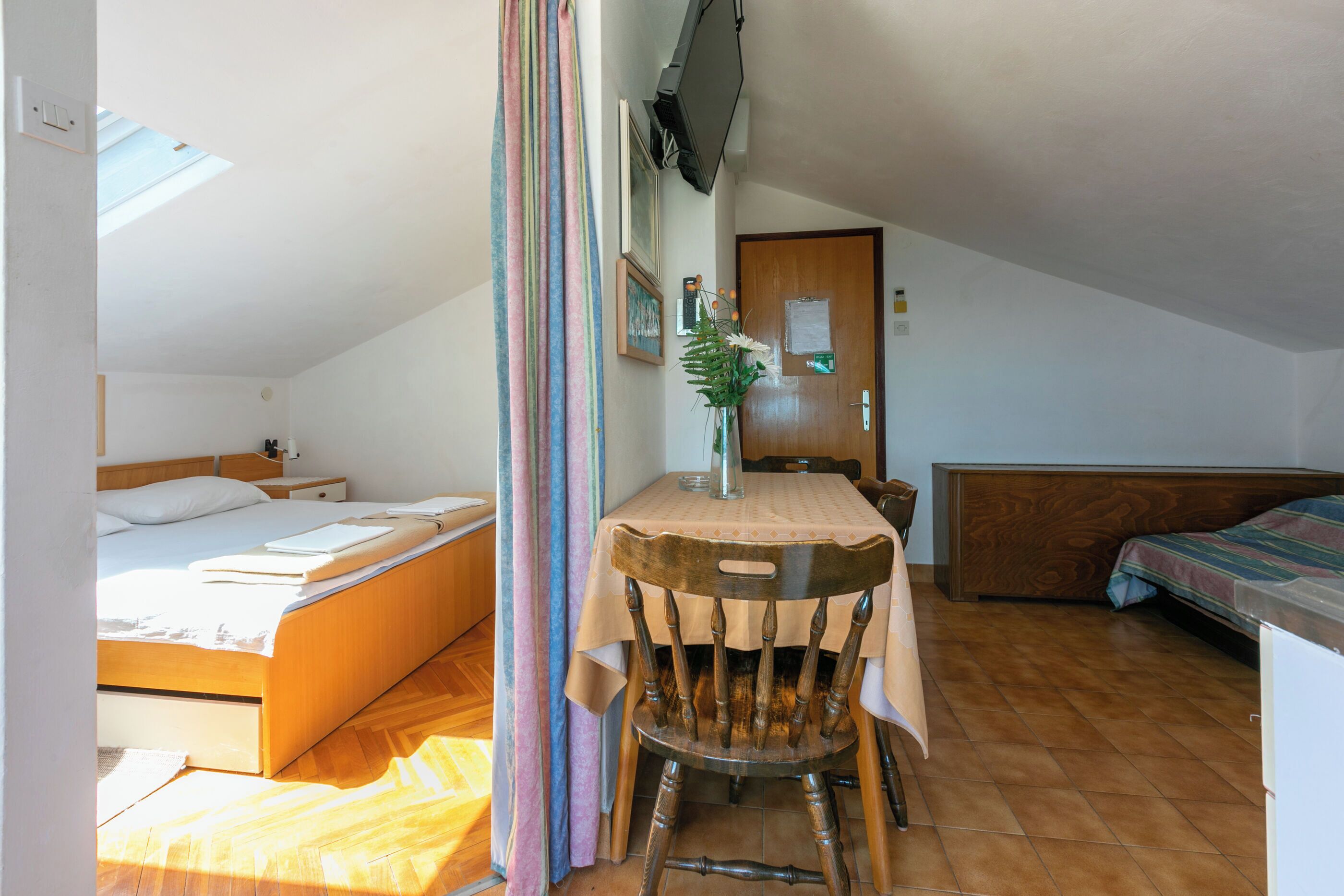 A small, cozy room with a bed, a wooden table with a chair, a small desk, and a TV mounted on the wall.