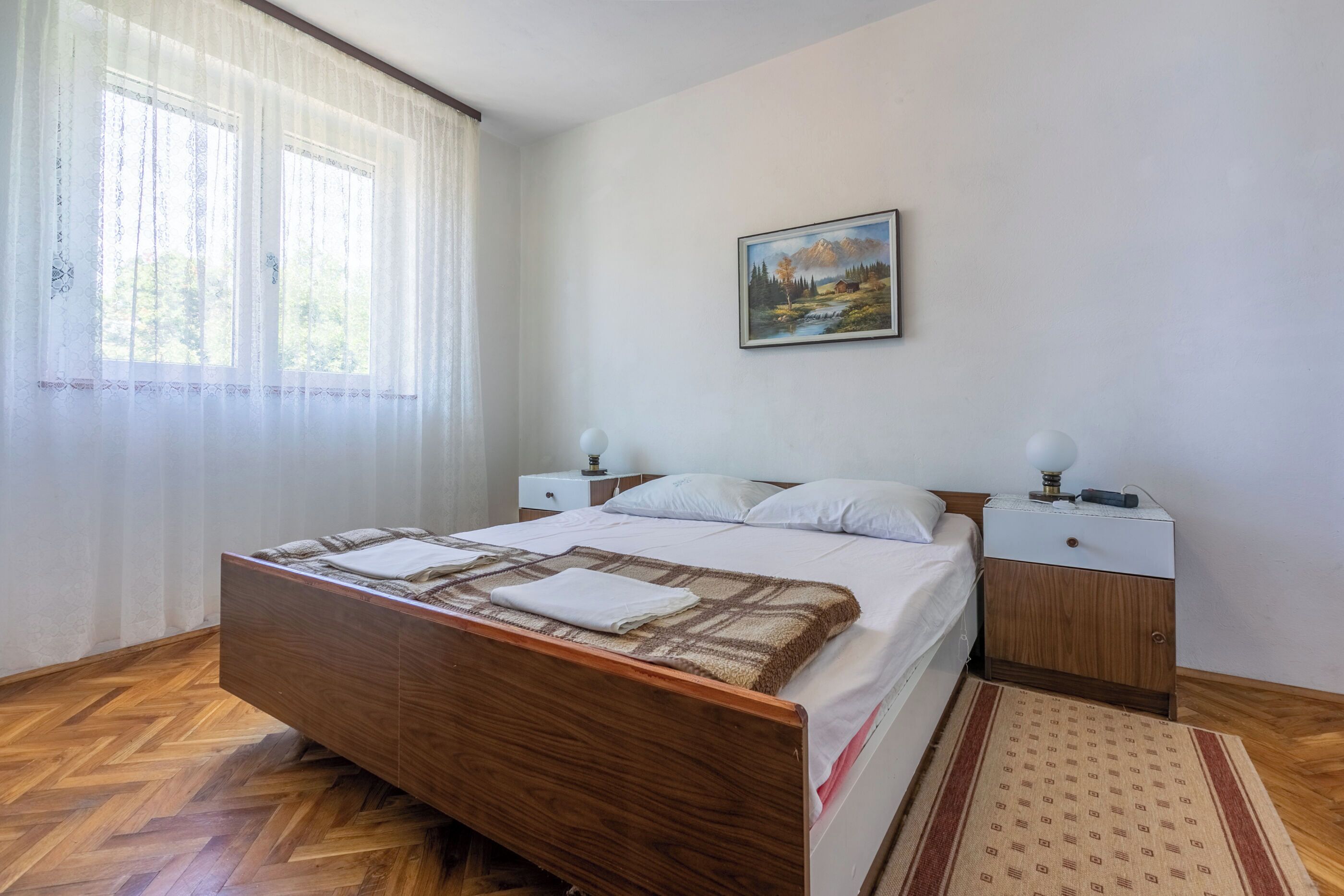A bedroom with a wooden bed, white bedding, a painting on the wall, and a nightstand with a lamp.