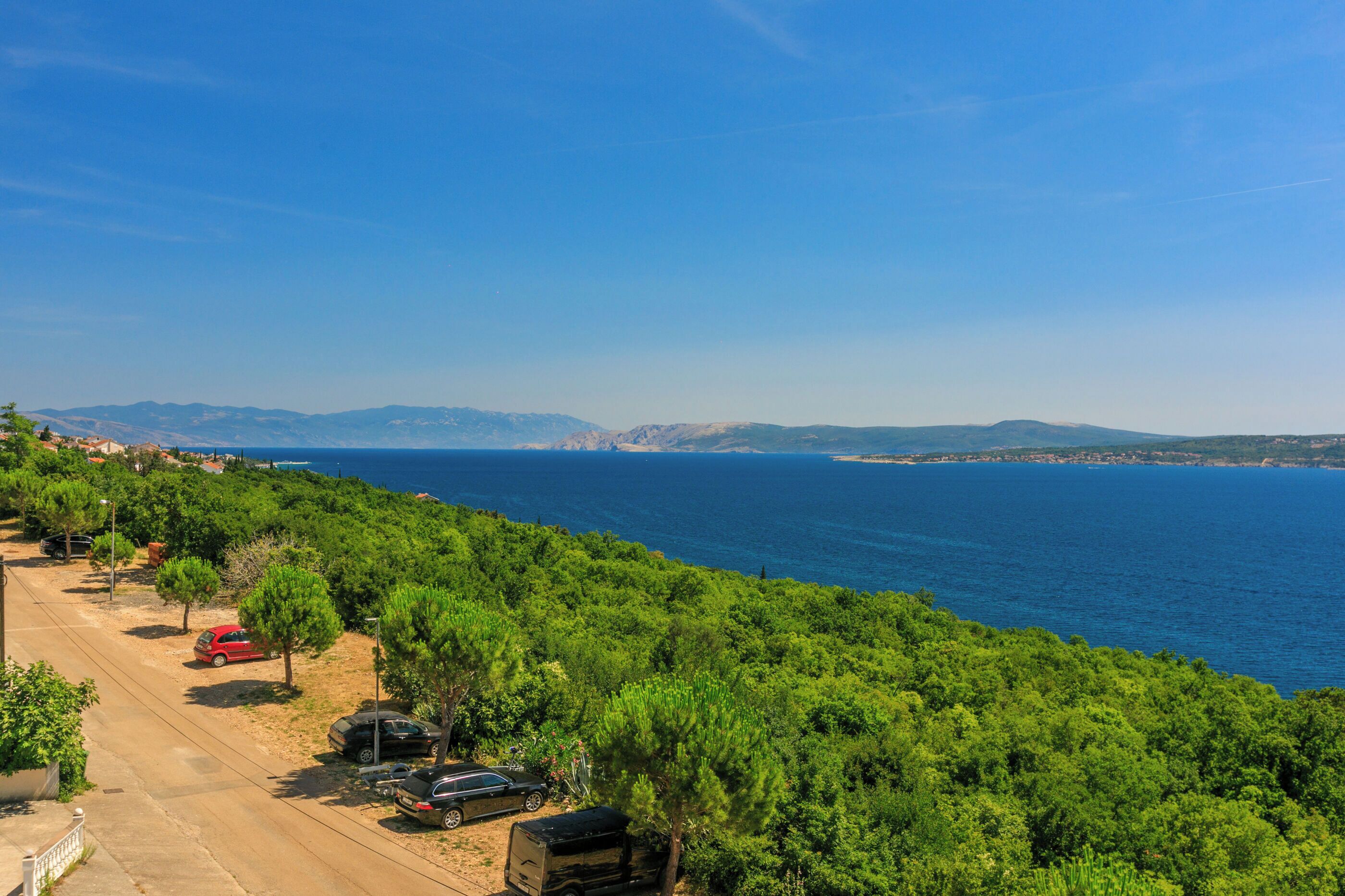 A coastal road with parked cars, lush greenery, and a view of the sea and distant mountains.