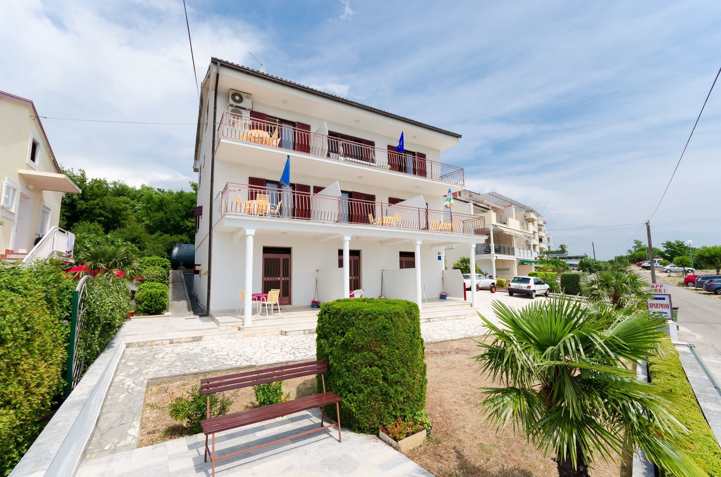 A multi-story residential building with balconies, a paved entrance, and a small garden area.
