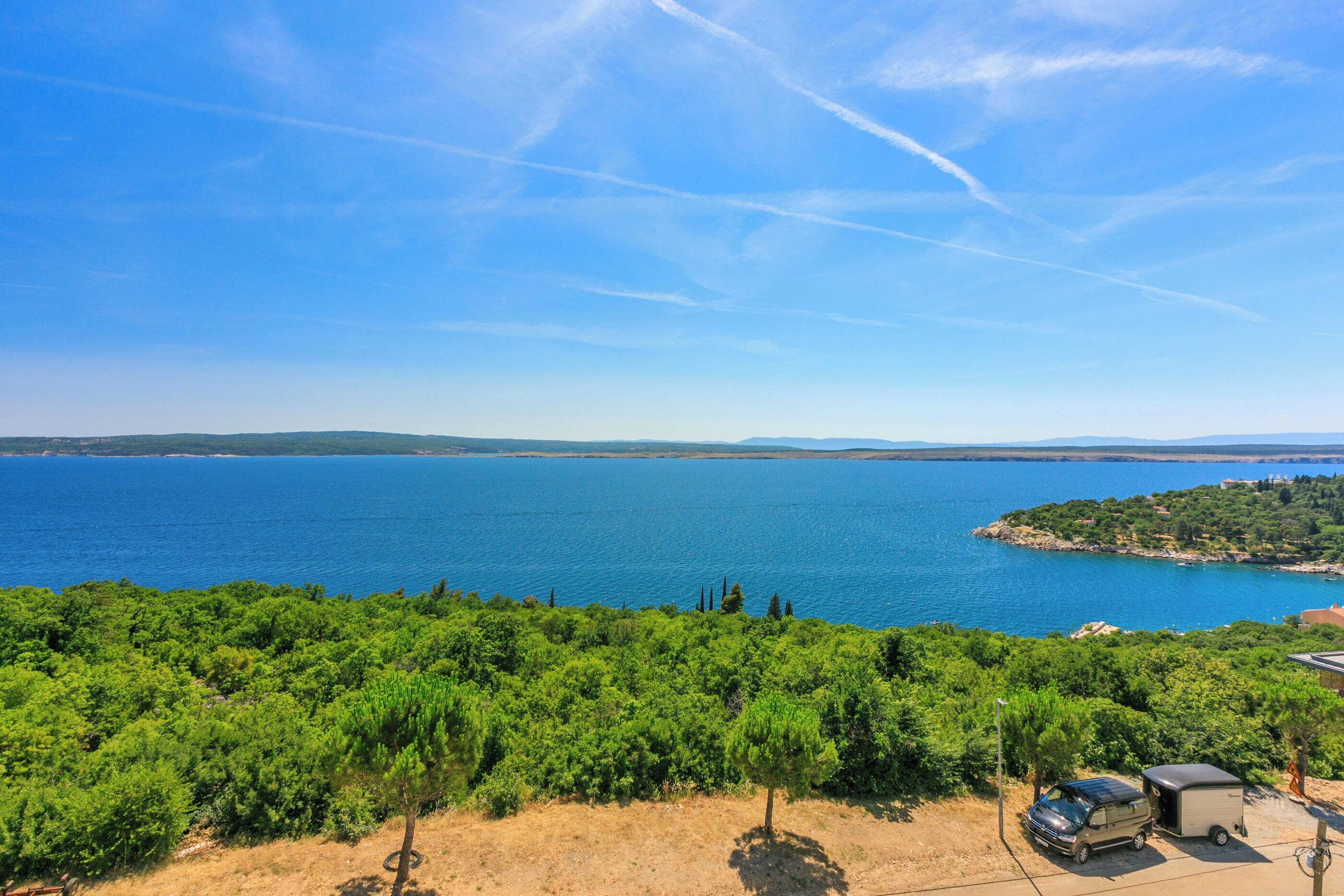 A coastal landscape with a clear view of the sea, a parking area with a car and a trailer, and dense greenery in the foreground.