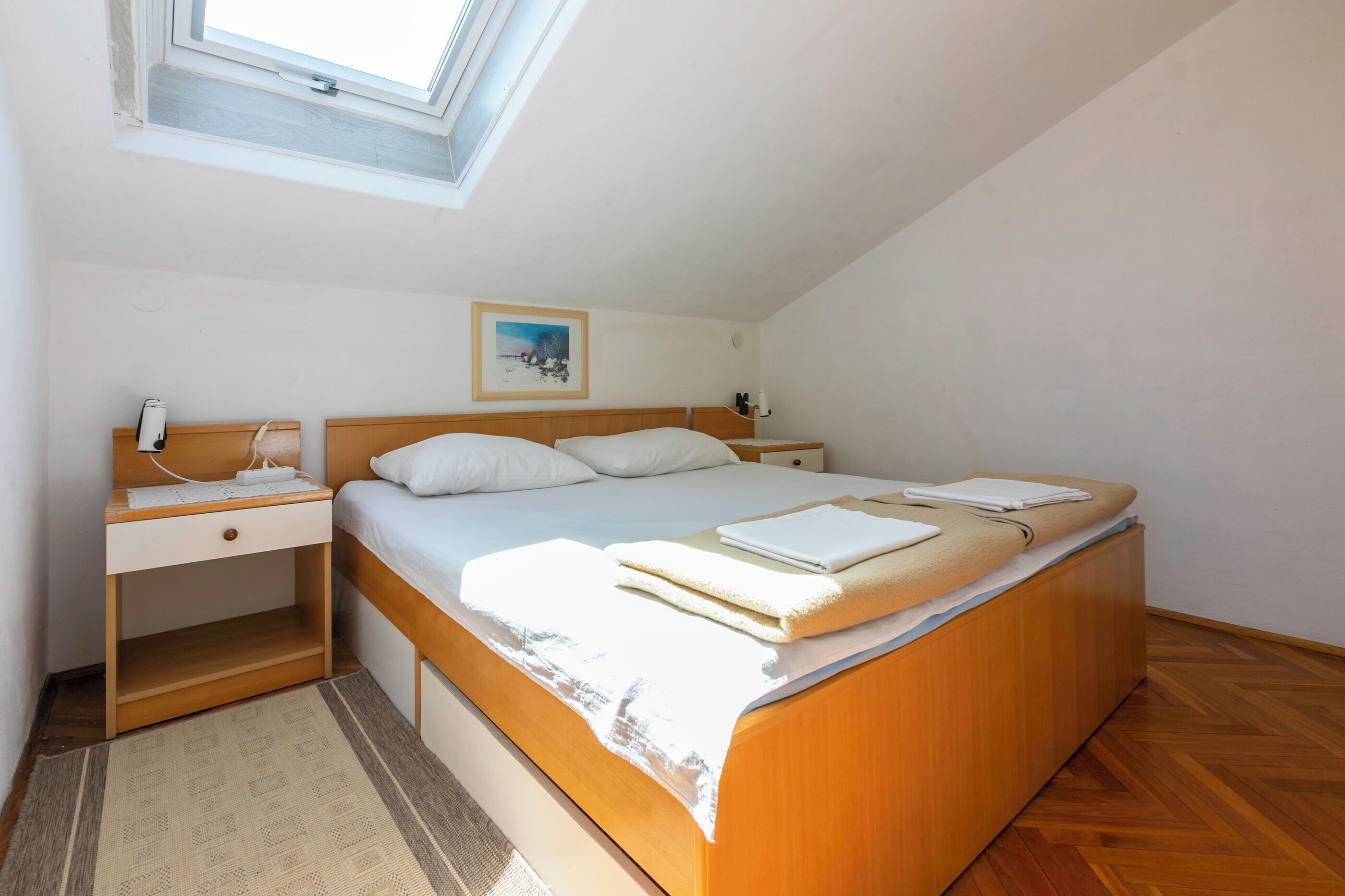 Een slaapkamer met een houten bed, een bureau met lamp, een klein nachtkastje, een schilderij aan de muur en een dakraam.