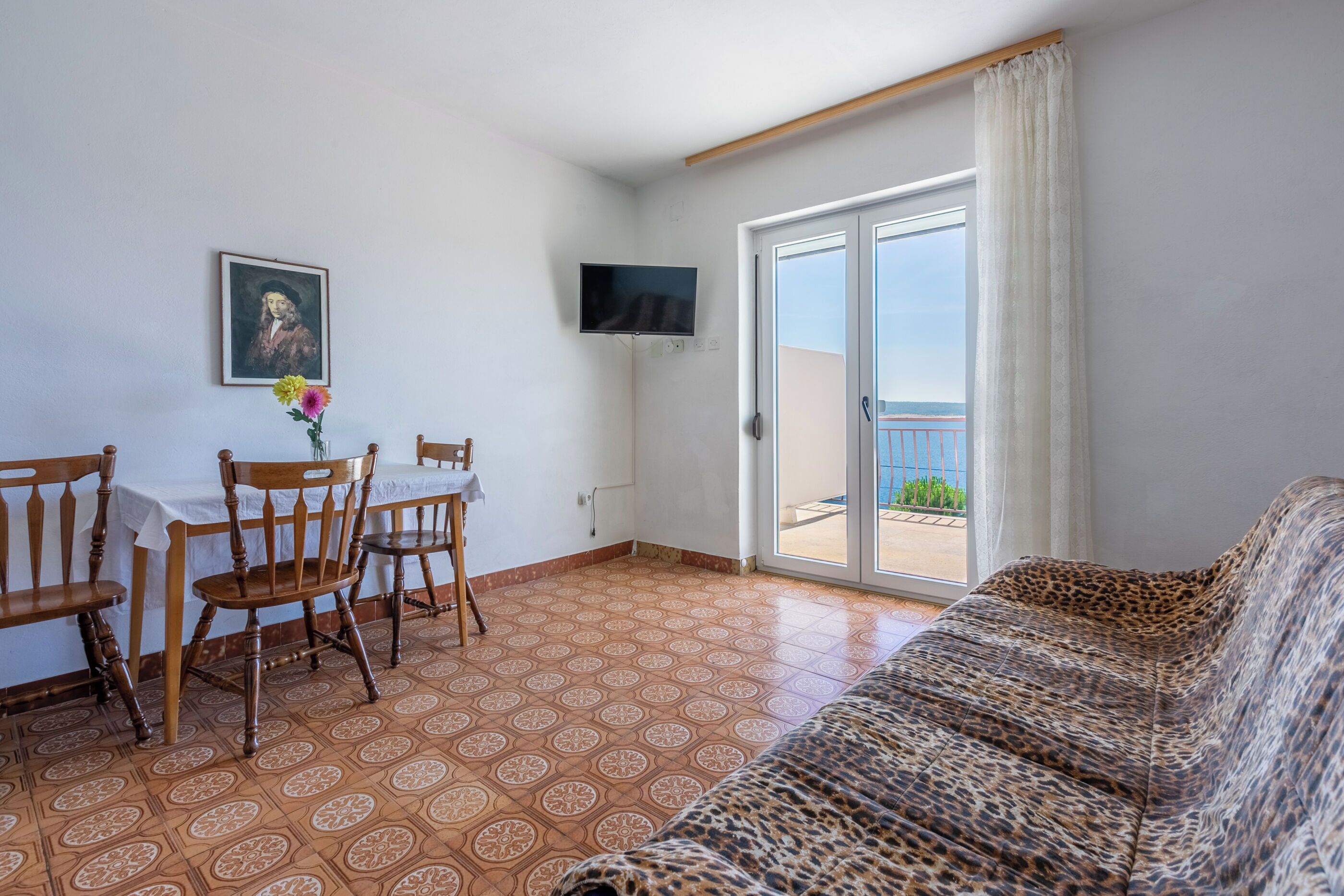 A living room with a sofa, a dining table with chairs, a TV mounted on the wall, and a large sliding glass door leading to a balcony.