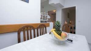 A dining table with a fruit bowl containing a pineapple, banana, and apple, and a wine glass. There are wooden chairs around the table. - Apartments Neno (Primosten)