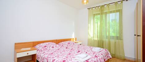 A bedroom with a wooden bed, a nightstand, a desk, and a window with sheer curtains.