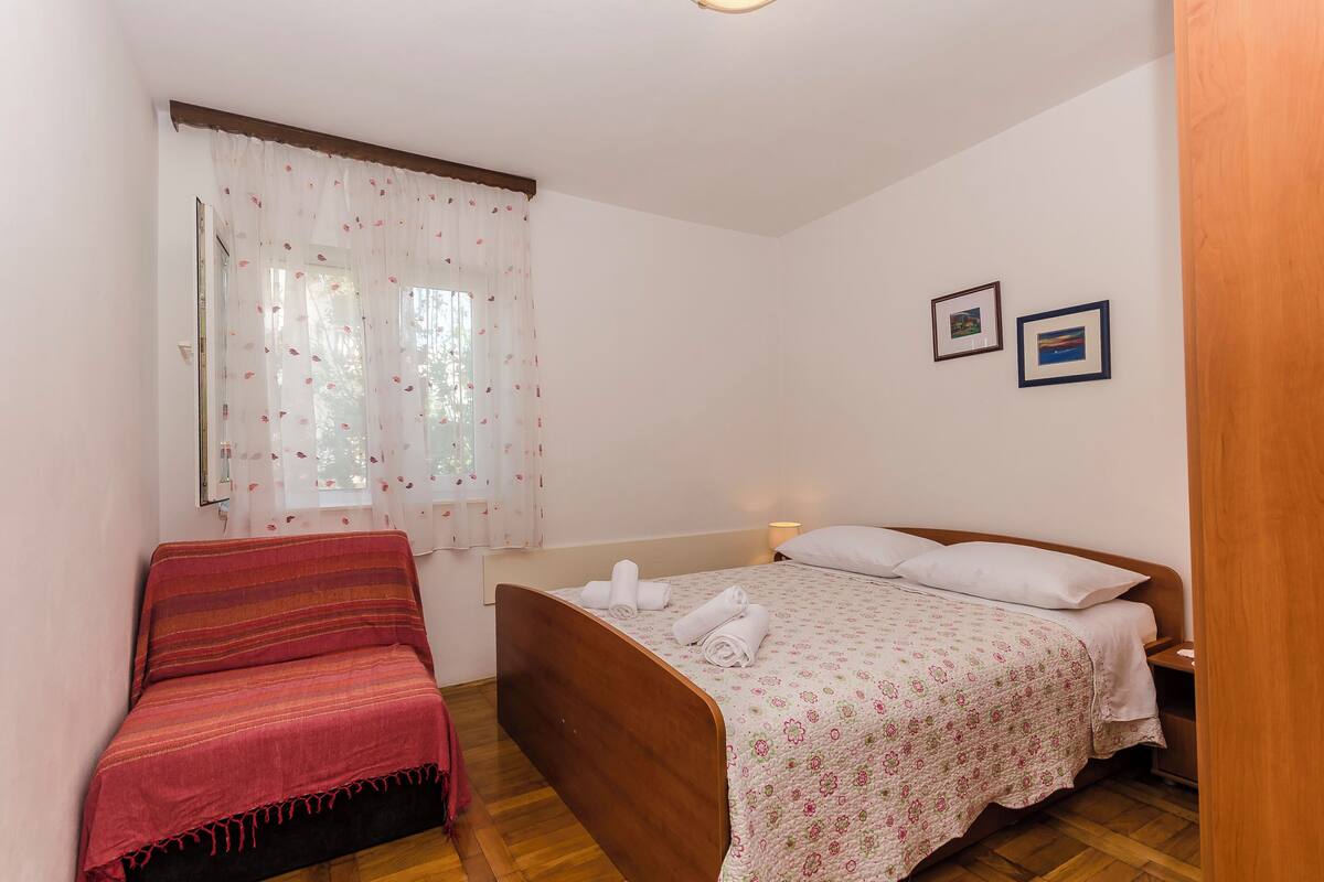 A bedroom with a bed, a chair, a window with curtains, and two framed pictures on the wall.