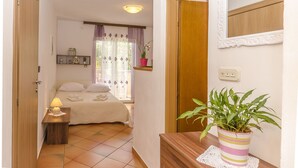 A bedroom with a bed, bedside table with a lamp, a window with curtains, a shelf with decorative items, and a potted plant. - Apartments Bella Vista (Srima)