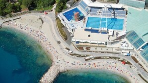 An aerial view of a coastal area with a swimming pool, beach, and adjacent buildings. - Apartments Marija (Rijeka)