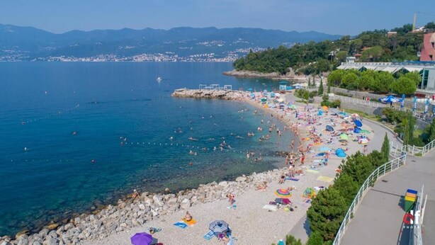 A coastal beach with clear water, rocky shore, and a view of distant mountains. - Apartments Marija (Rijeka)