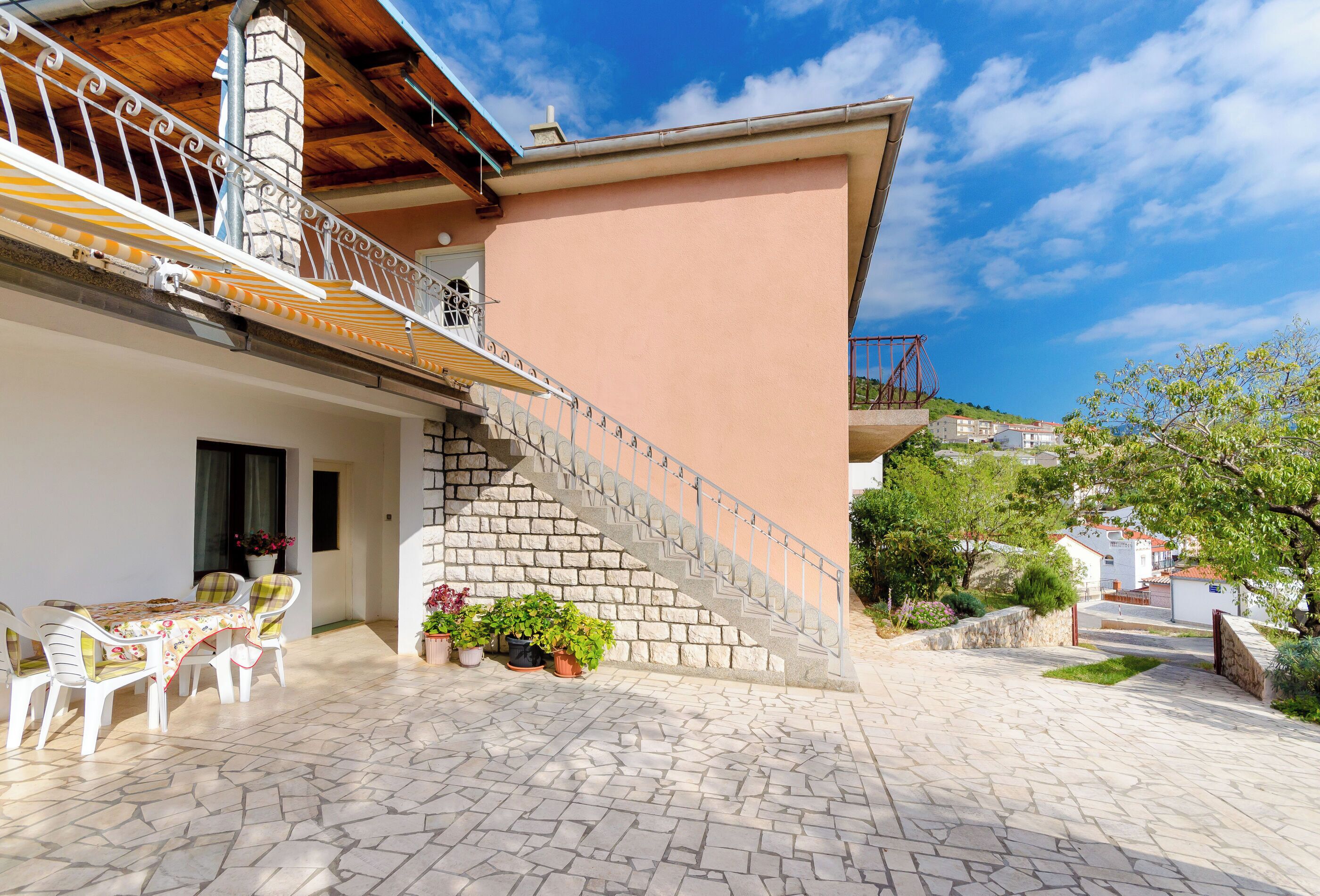 A stone-floored patio with outdoor furniture, a staircase with metal railings, and a building with a pink wall and a chimney.
