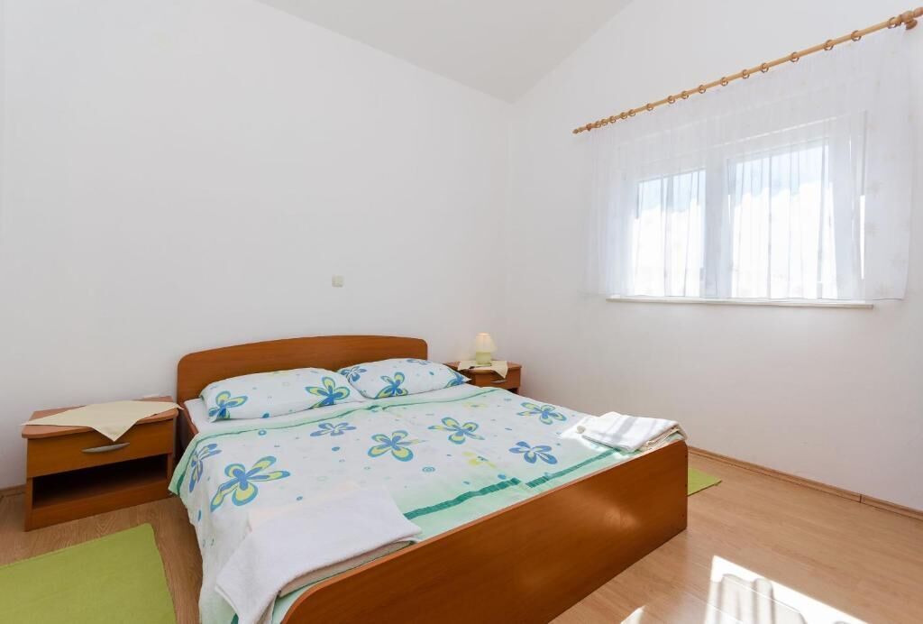 Een slaapkamer met een houten bed, een nachtkastje, een lamp en een raam met dunne gordijnen.