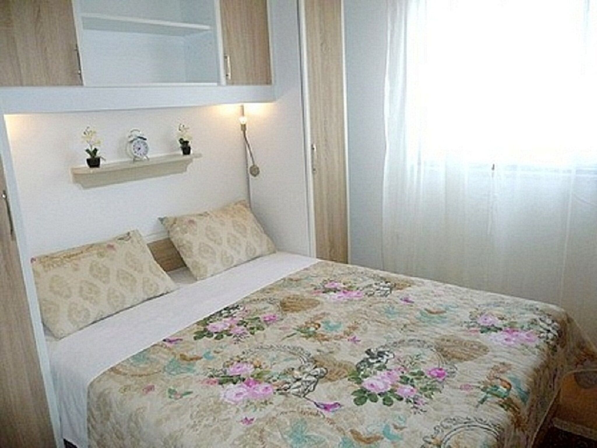 A bedroom with a floral bedspread, a bedside table with a lamp, and a shelf with decorative items.