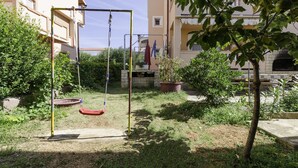 A playground with a swing set and a tire swing, surrounded by greenery and a building with balconies. - Apartments Biserka (Barbat)