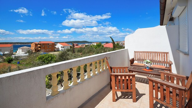 A balcony with wooden furniture, a view of the sea, and buildings in the background. - Apartments Biserka (Barbat)