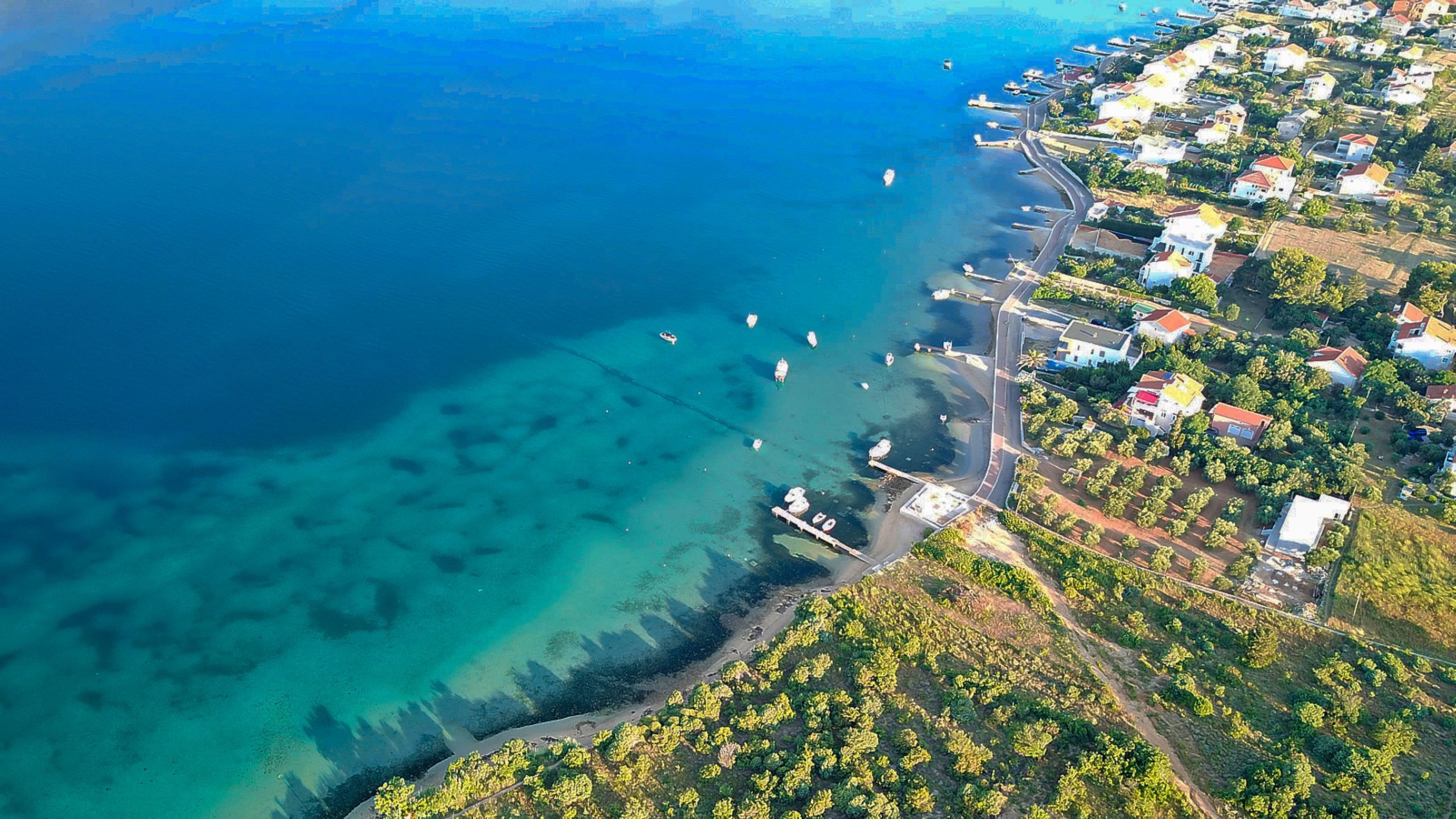 An aerial view of a coastal town with clear blue waters, a sandy beach, and residential buildings along the shore.
