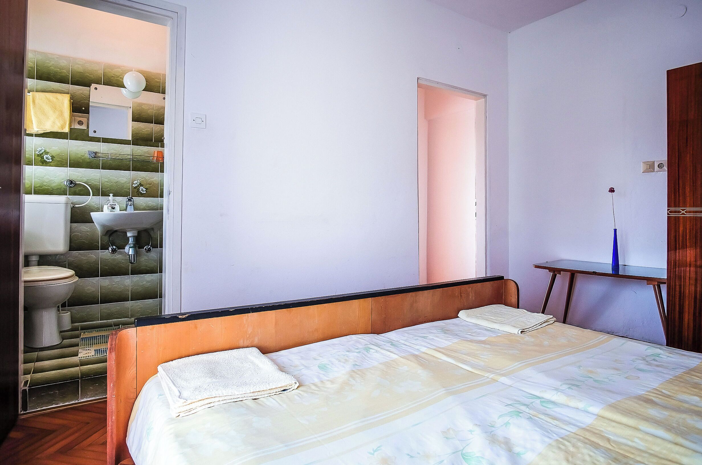 A bedroom with a bed, a bedside table, a wooden headboard, and a bathroom visible through an open door.