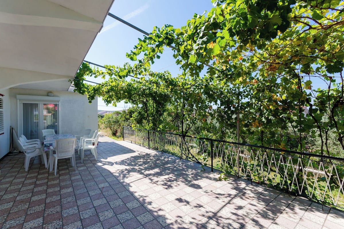 A covered patio area with a table and chairs, a railing, and a view of a tree-lined path.