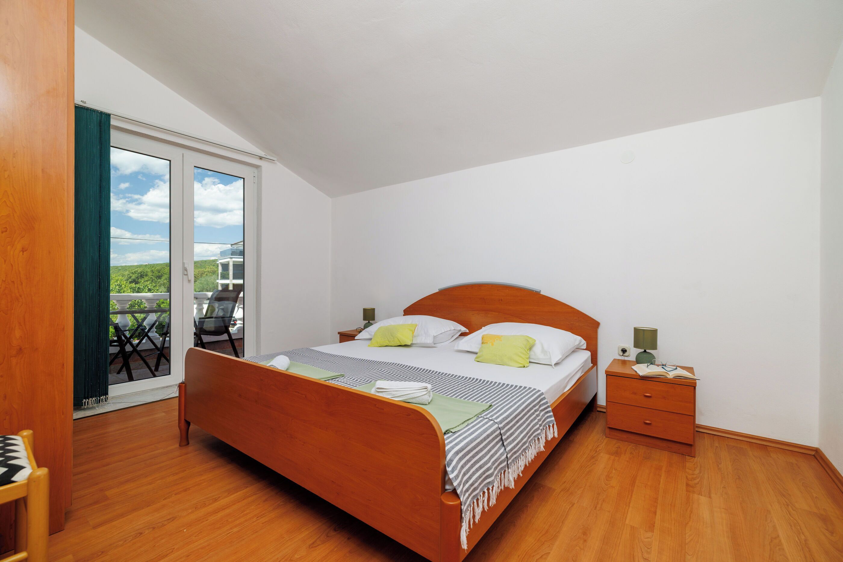 Ein Schlafzimmer mit einem hölzernen Bett, einem Nachttisch, einem Sessel und einer Glastür, die auf einen Balkon führt.