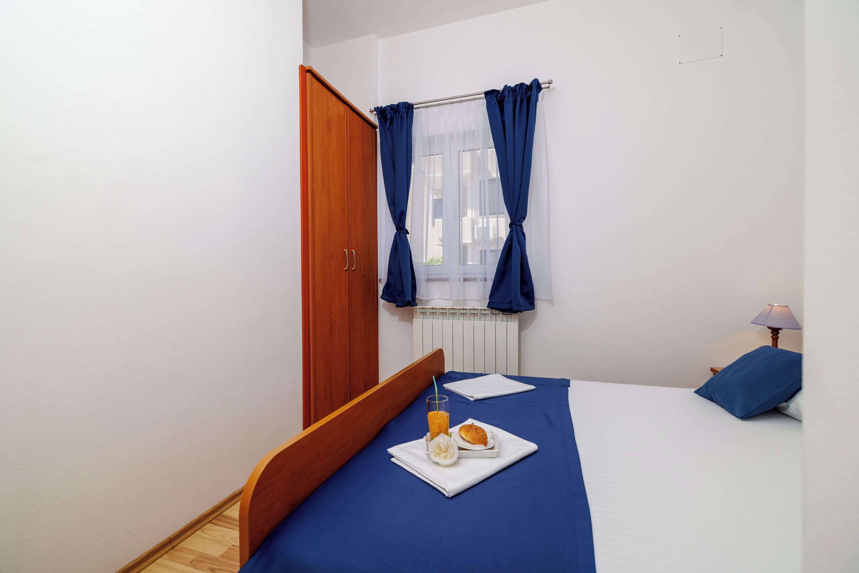 Ein Schlafzimmer mit einem hölzernen Bett, einem Fenster mit blauen Vorhängen, einer Nachttischlampe und einem Frühstücksbrett mit Orangensaft und einem Gebäckstück.
