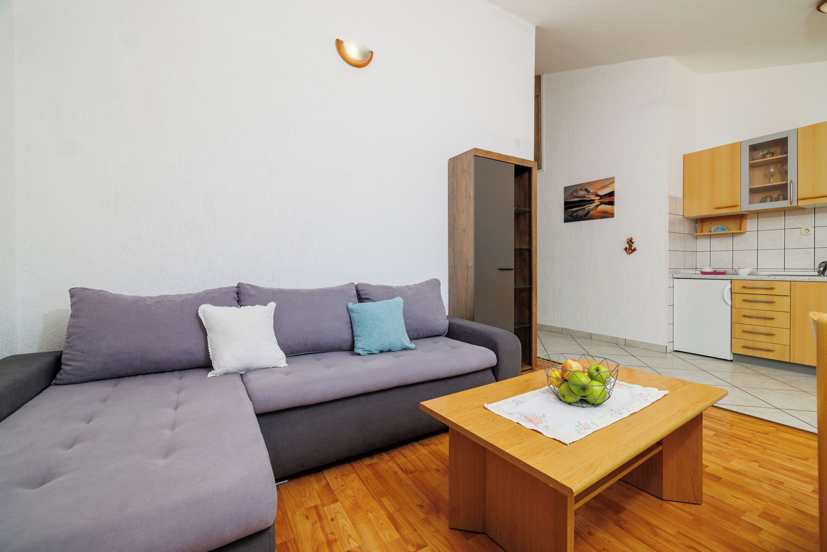 A modern living room with a grey sofa, a wooden coffee table with a fruit bowl, and a kitchen in the background.