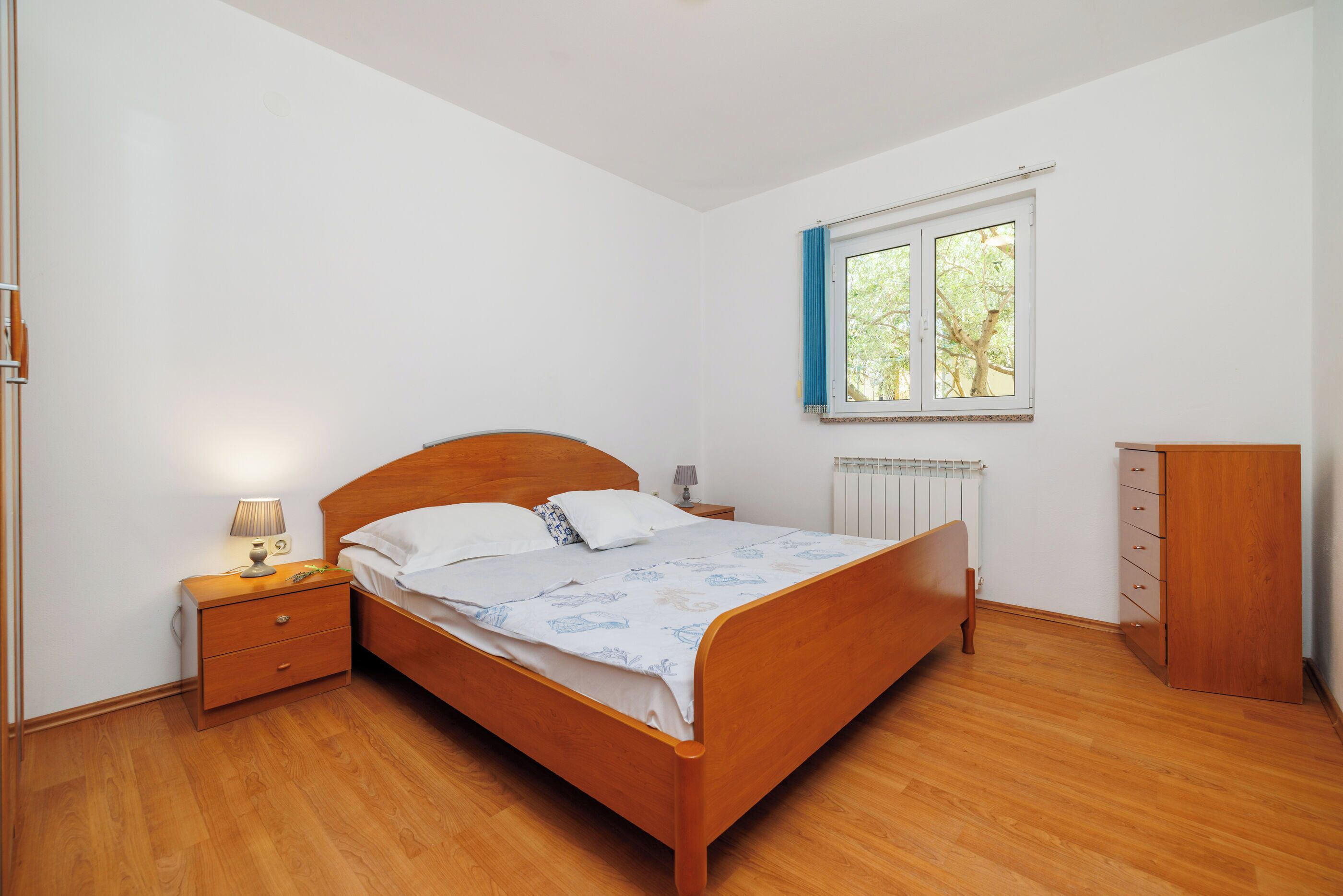 A bedroom with a wooden bed, a nightstand with a lamp, a dresser, and a window with curtains.