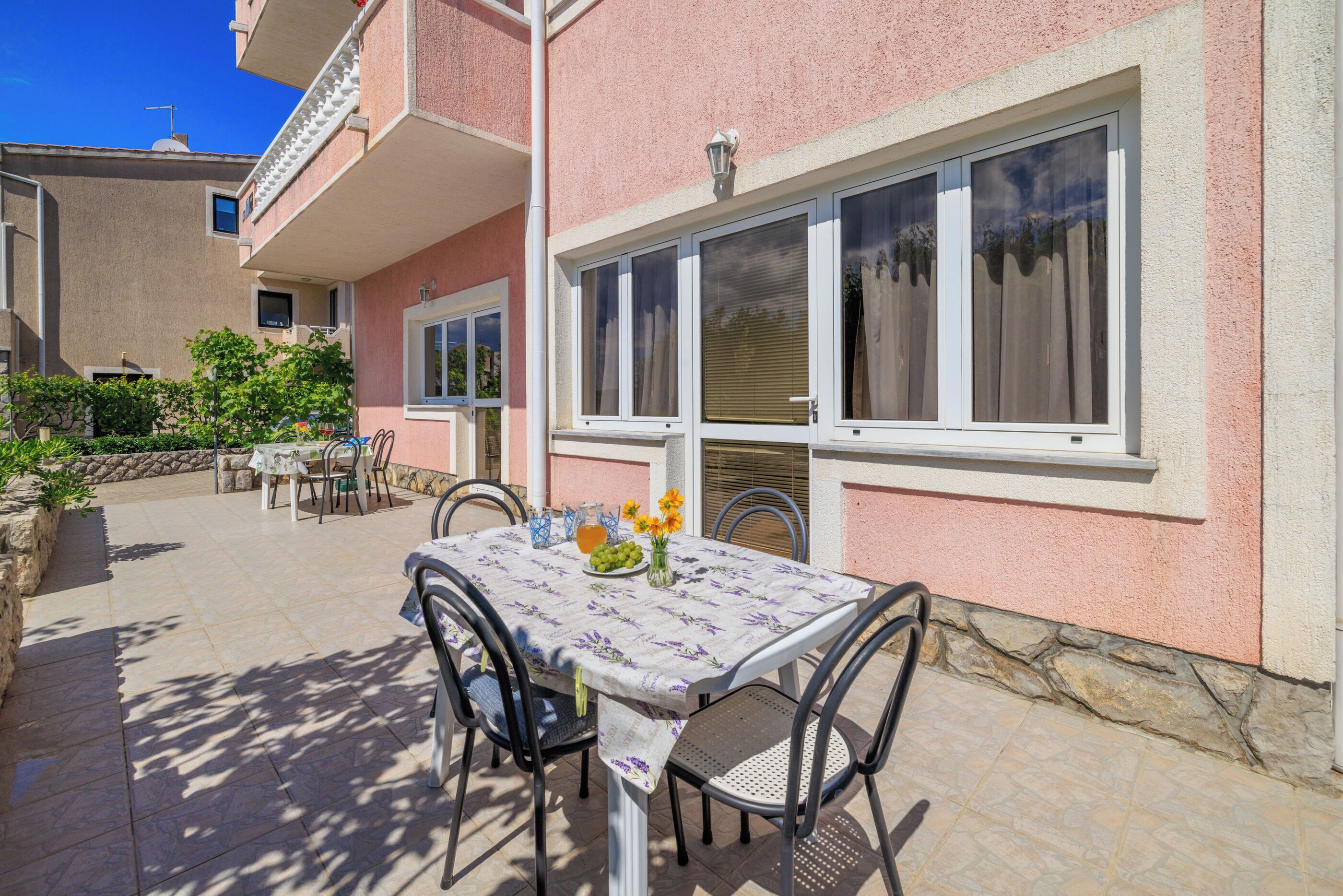 Un patio al aire libre con una mesa lista para comer, rodeada de sillas y una ventana con cortinas.