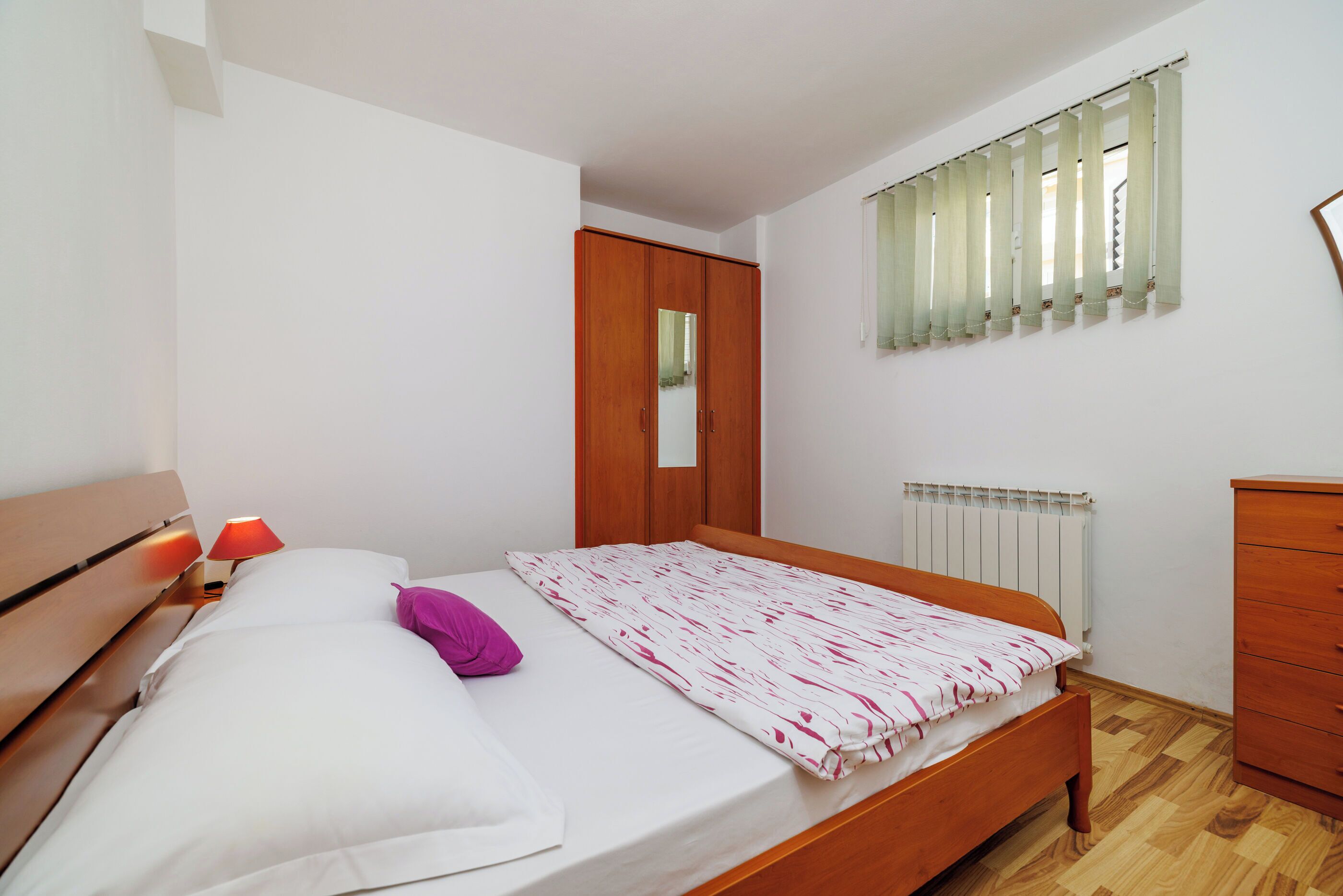 Ein Schlafzimmer mit einem hölzernen Bett, einem Nachttisch mit Lampe, einem Kleiderschrank und einem Fenster mit Jalousien.