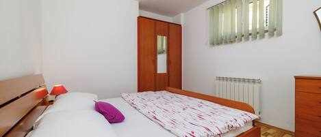 A bedroom with a wooden bed, a nightstand with a lamp, a wardrobe, and a window with blinds.