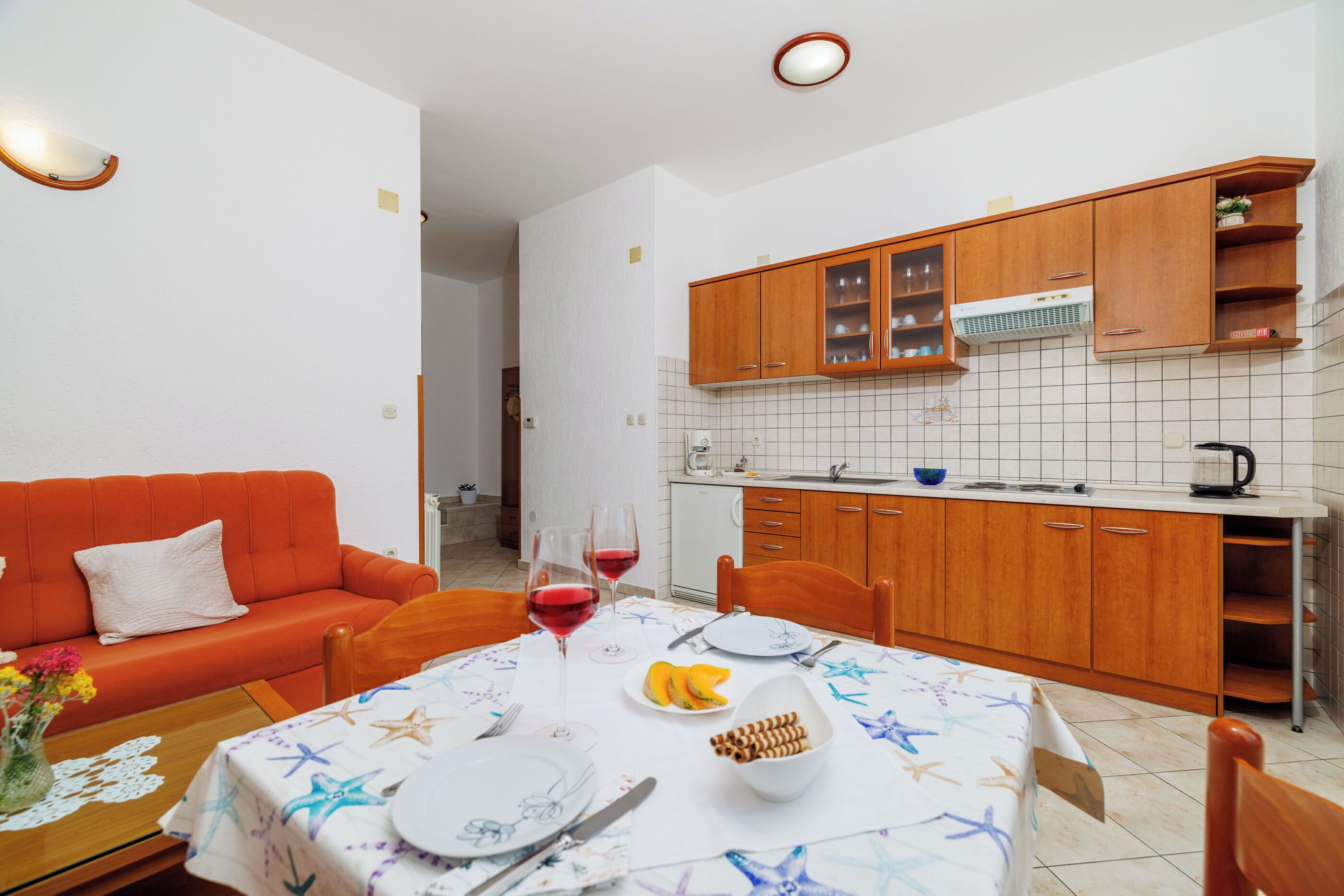 A compact kitchen with wooden cabinets, a dining table set with plates, glasses, and a bowl of snacks.