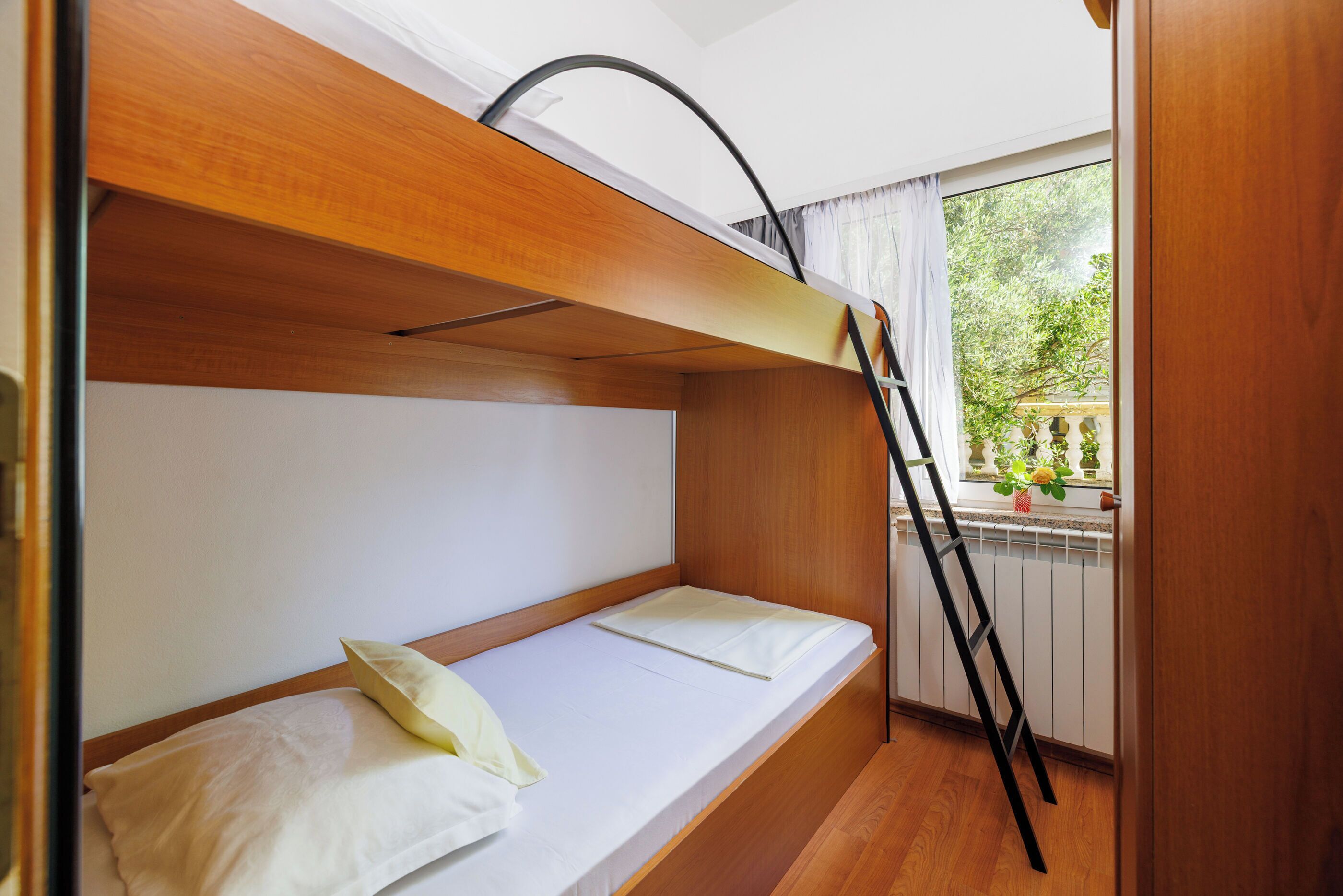 Una cama litera con cabecera de madera, una escalera y una ventana que da a vistas de vegetación.