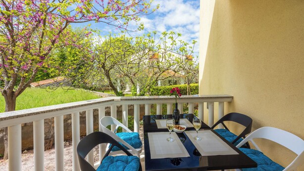 A balcony with a table and chairs, a view of a garden with blooming trees, and a building in the background.