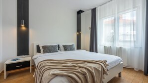 A modern bedroom with a large bed, a bedside table, a wall-mounted light, and a window with curtains.