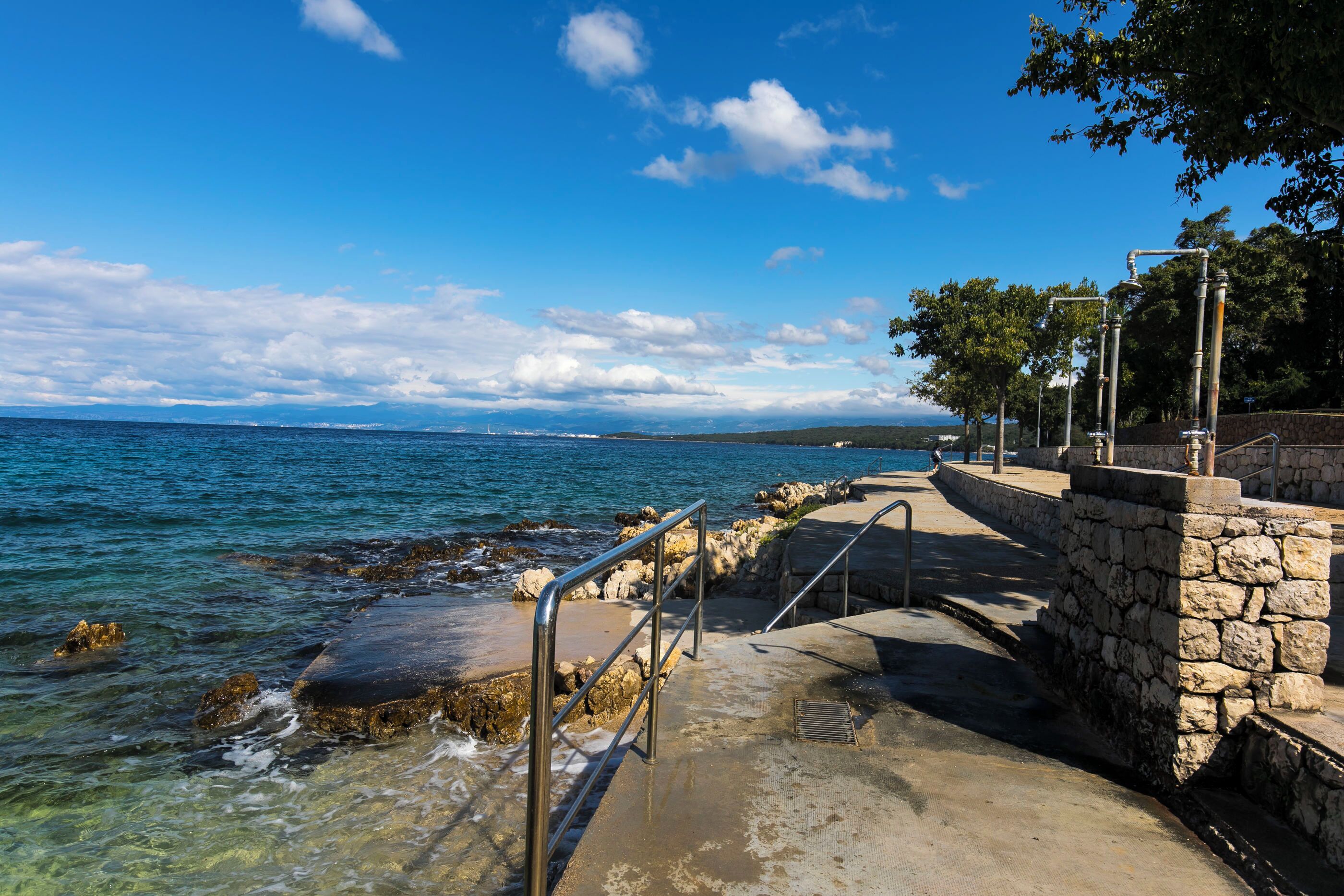 Une promenade en bord de mer, bordée de murs en pierre et de rampes métalliques, qui mène à la mer.